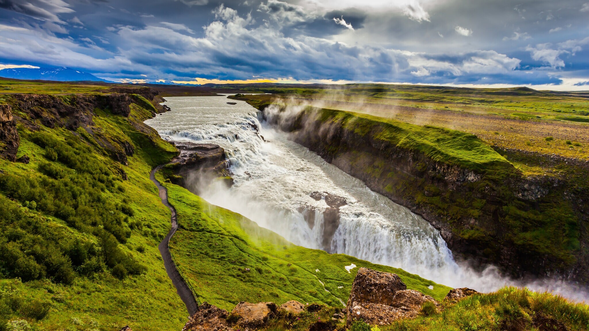 Breiter Wasserfall stürzt in zwei Stufen in eine tiefe Schlucht, umgeben von grünen Wiesen und bewölktem Himmel