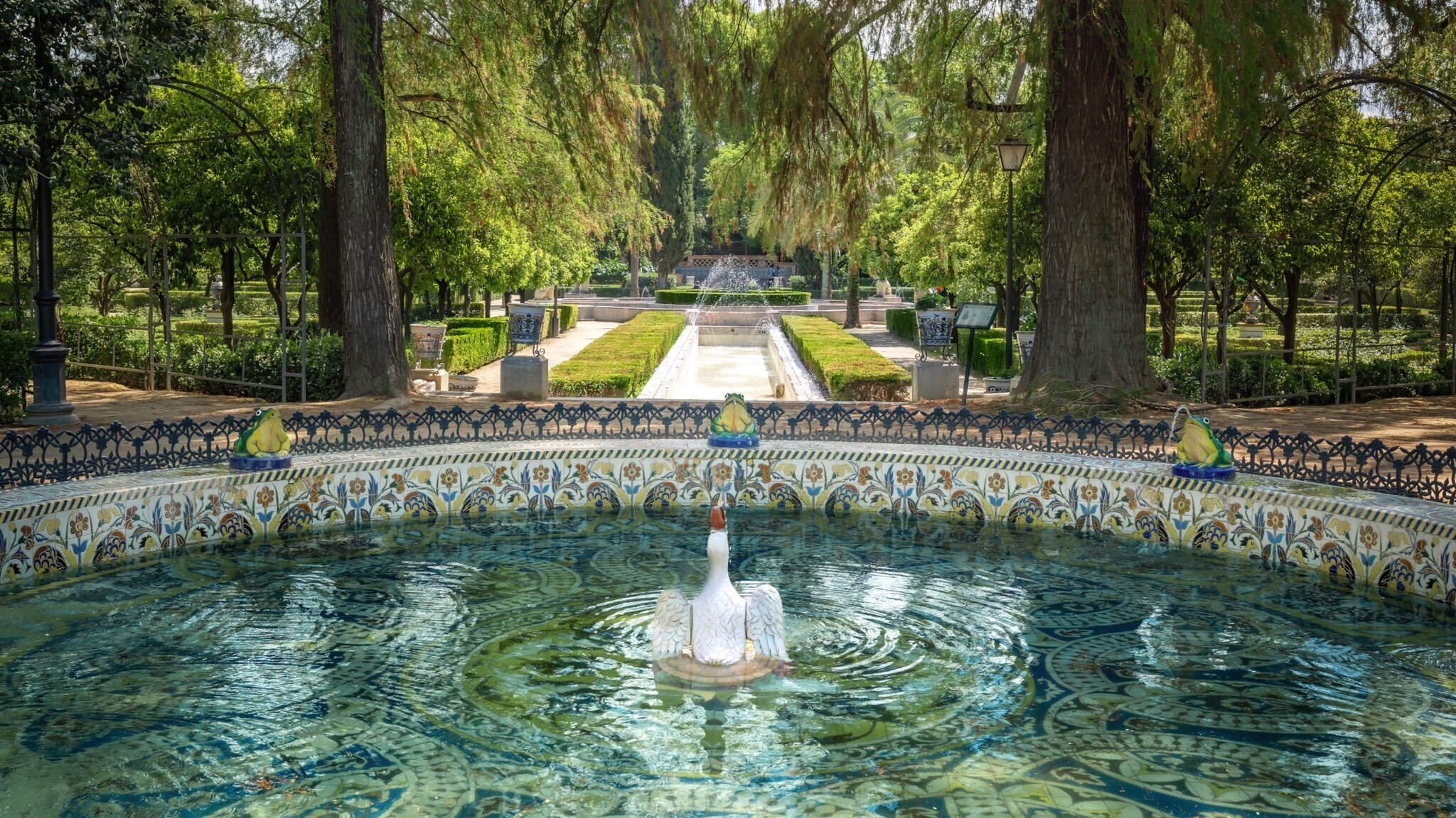 Verzierter Brunnen mit weißer Schwan-Skulptur in der Mitte, umgeben von Froschfiguren in einem Park mit Bäumen