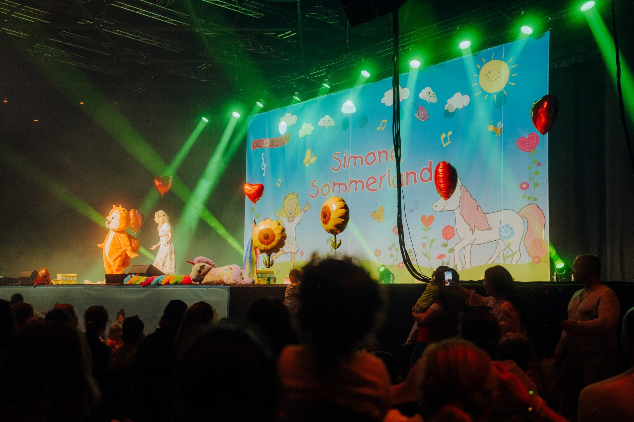 Simone Sommerland steht auf einer Bühne voller Luftballons, neben ihr eine Person im Eichhörnchen-Kostüm, vor ihr Publikum. Simone Sommerland steht auf einer Bühne voller Luftballons, neben ihr eine Person im Eichhörnchen-Kostüm, vor ihr Publikum.
