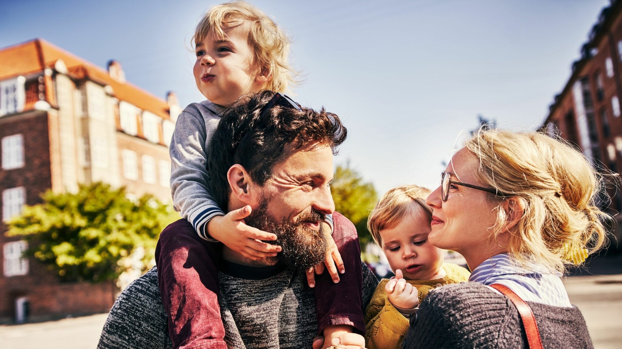 Vater trägt lachendes Kleinkind auf Schultern, Mutter mit Brille schaut liebevoll, während sie ein weiteres Kleinkind im Arm hat. Vater trägt lachendes Kleinkind auf Schultern, Mutter mit Brille schaut liebevoll, während sie ein weiteres Kleinkind im Arm hat.