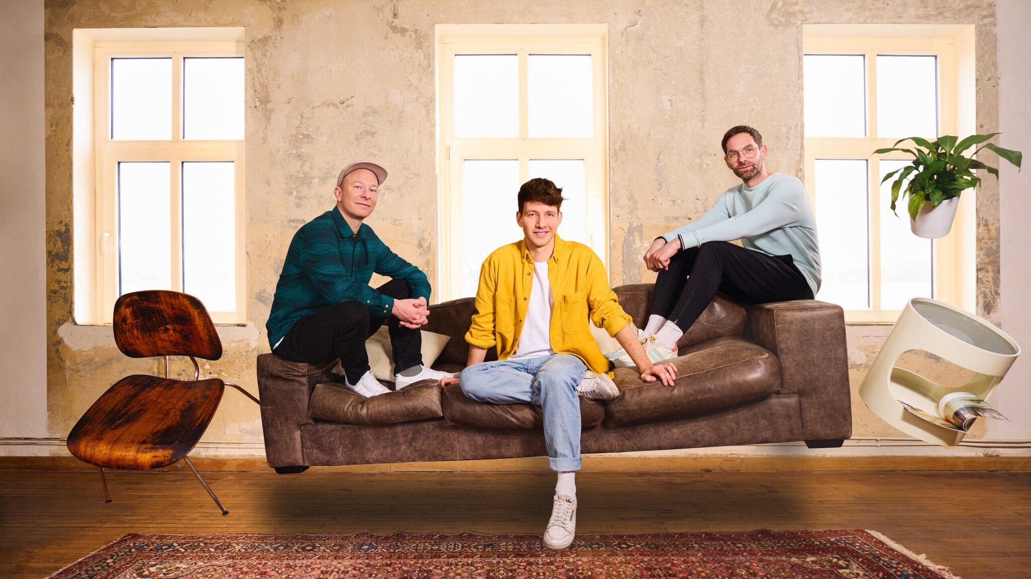 Drei Männer in lässiger Kleidung sitzen auf einer schwebenden braunen Ledercouch vor einer Wand mit drei Fenstern, daneben eine schwebende Pflanze und ein schwebender Stuhl. Drei Männer in lässiger Kleidung sitzen auf einer schwebenden braunen Ledercouch vor einer Wand mit drei Fenstern, daneben eine schwebende Pflanze und ein schwebender Stuhl.