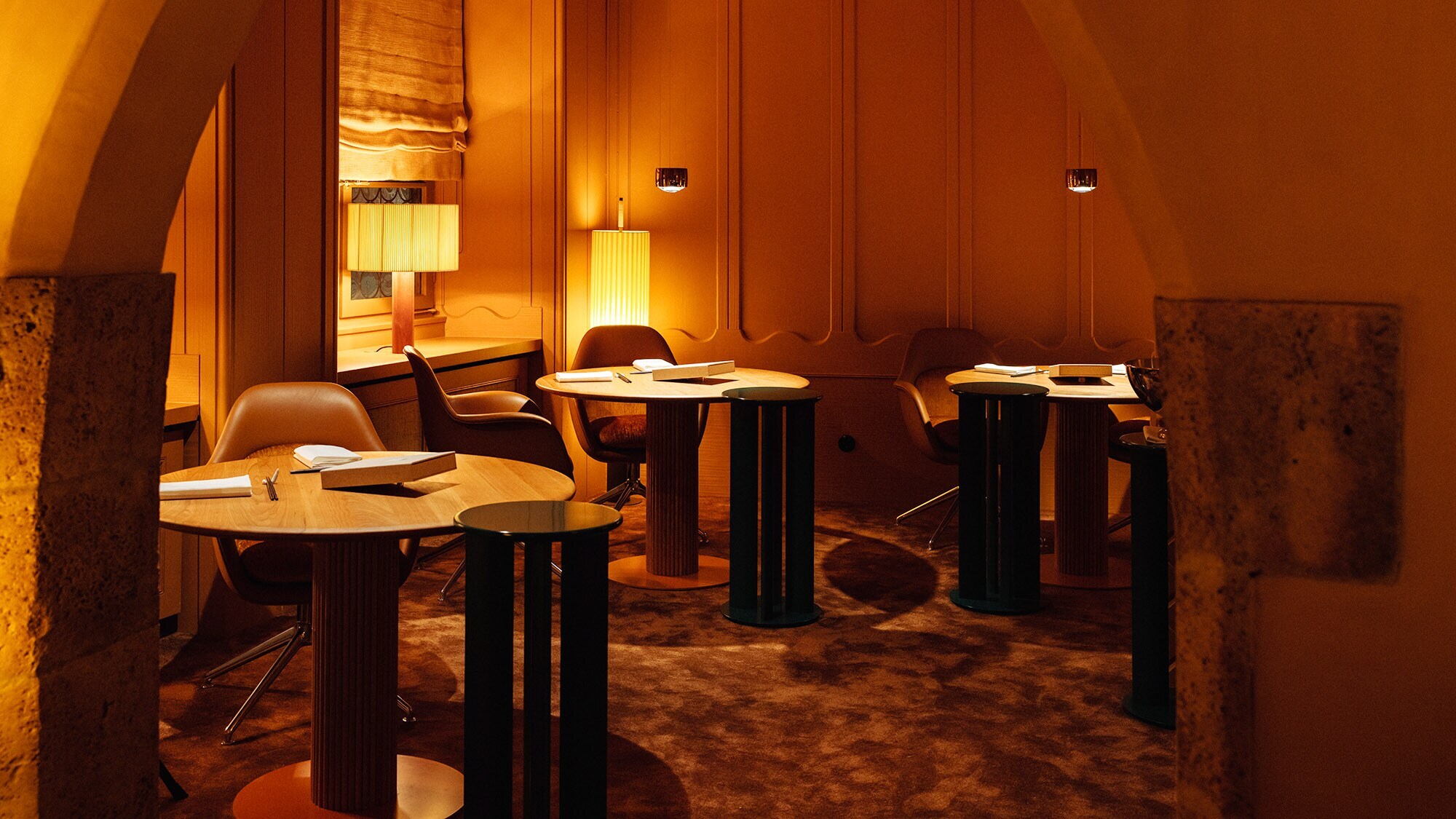 Stilvoll eingerichteter Gastraum mit runden Holztischen auf Teppichboden bei gedimmter, orangefarbener Beleuchtung.