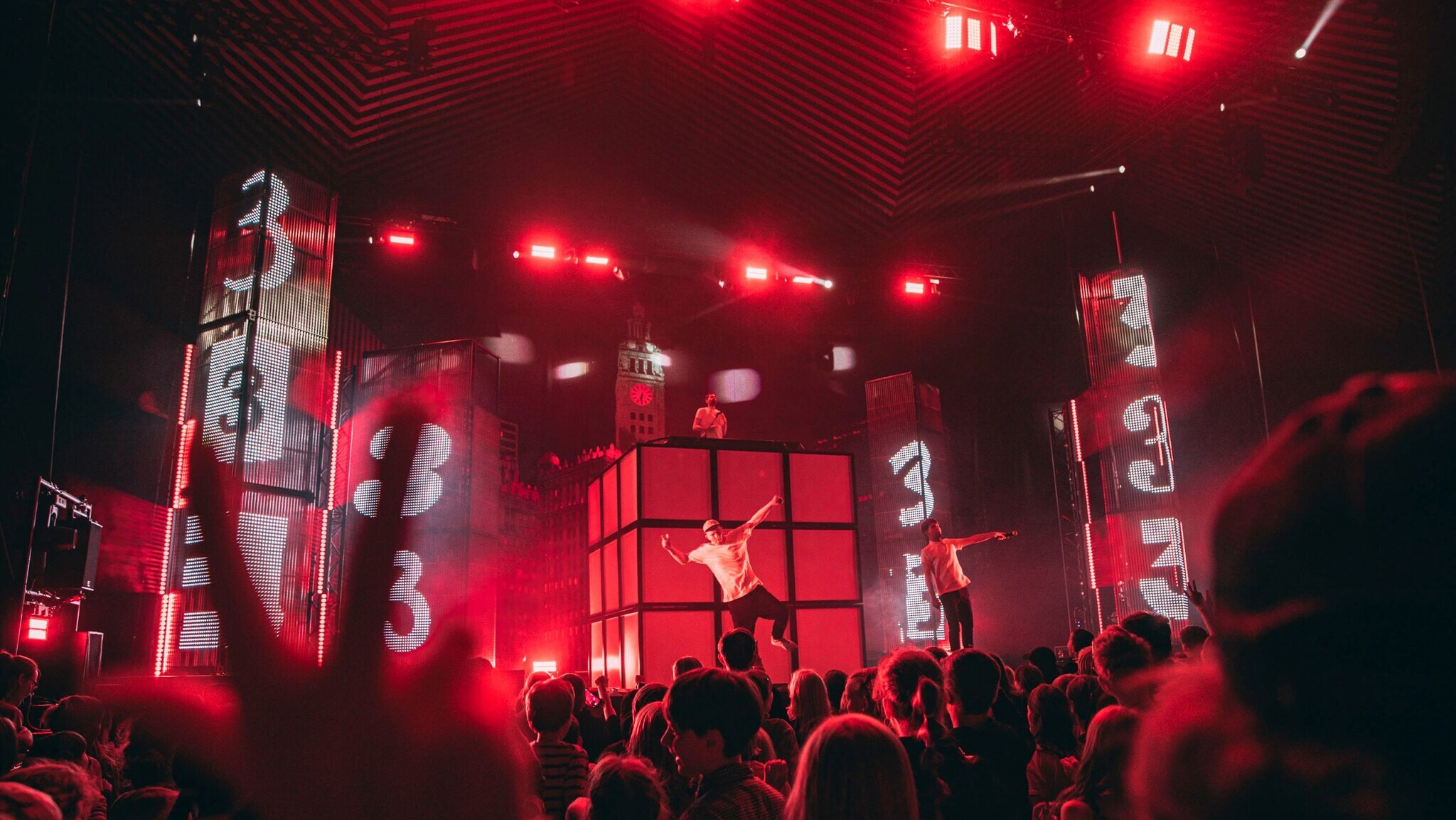 Musiker auf Bühne mit roter Beleuchtung und großen Bildschirmen, die die Zahl 3 zeigen, davor ist Publikum zu sehen.