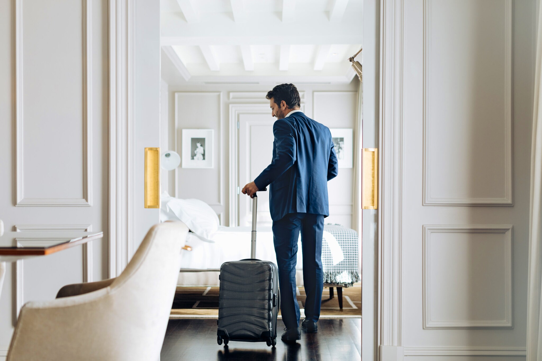 Mann im Anzug und einem Rollkoffer in der Hand betritt eine helle luxuriöse Hotel-Suite.