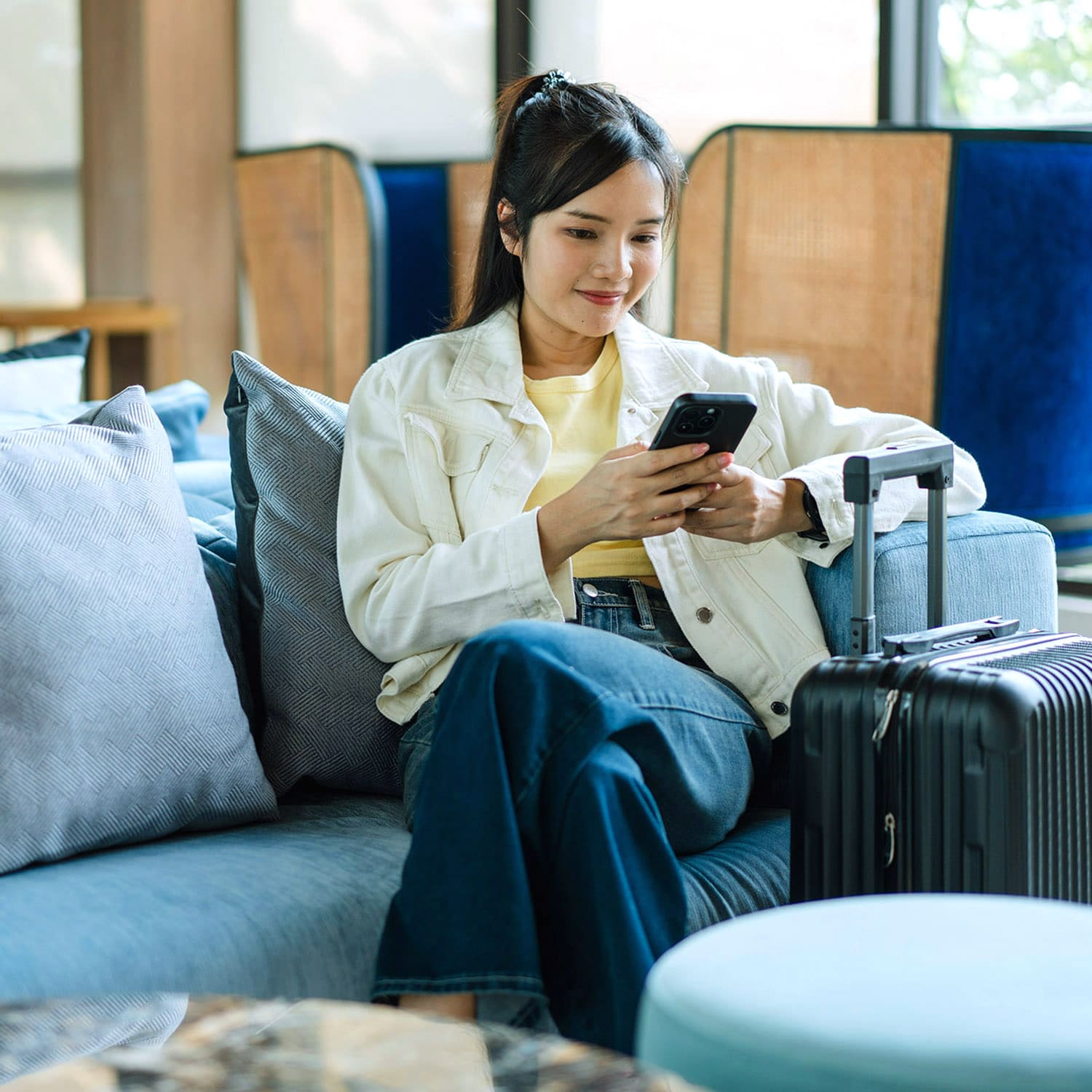 Eine junge asiatische Frau sitzt iauf einem Sessel in einer Lounge und schaut auf ihr Smartphone, neben ihr steht ein Hartschalenkoffer.