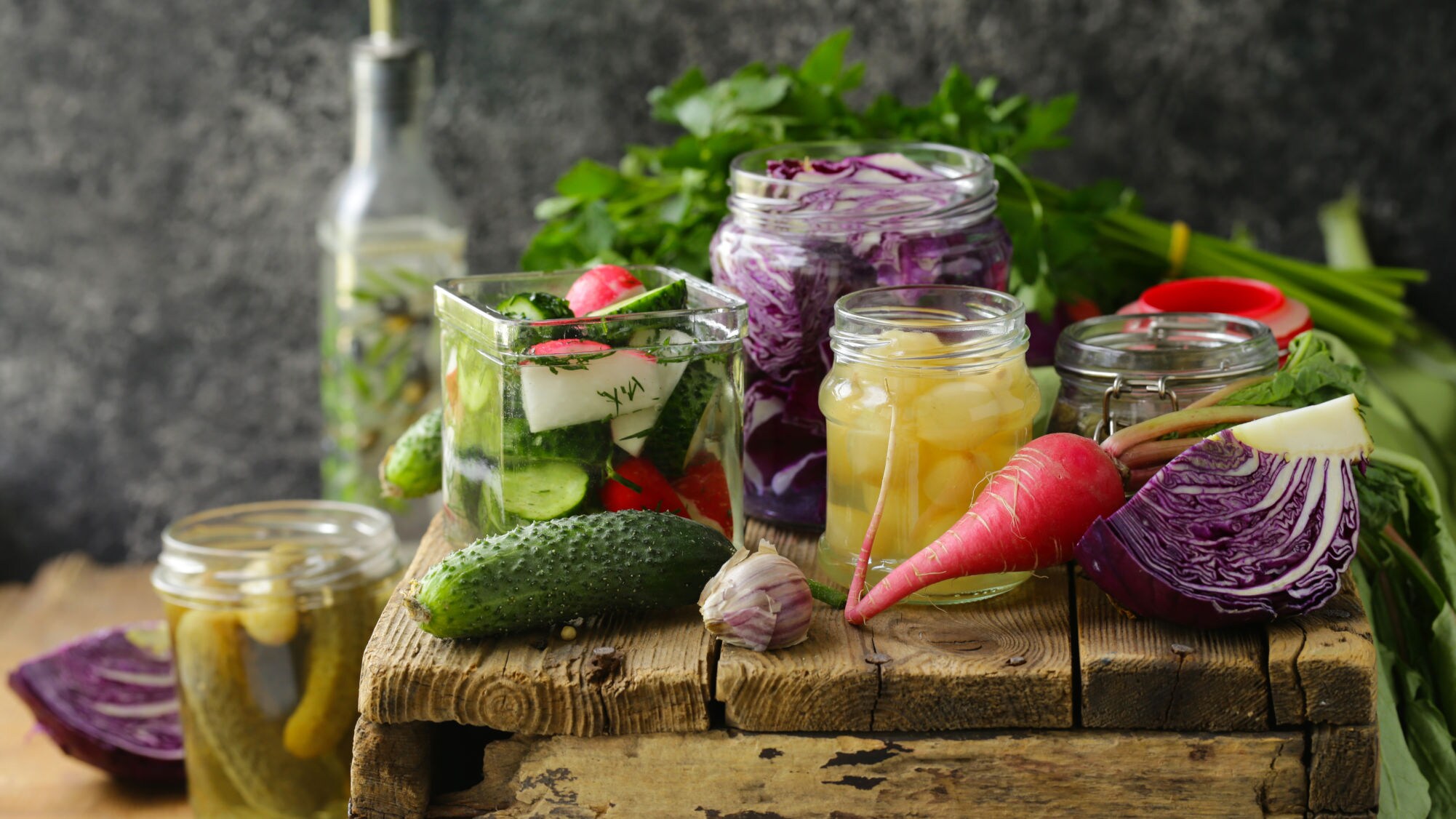 Verschiedene eingelegte Gemüse in Gläsern, darunter Gurken, Rotkohl und Zwiebeln, auf einem rustikalen Holztisch mit frischem Gemüse im Hintergrund.