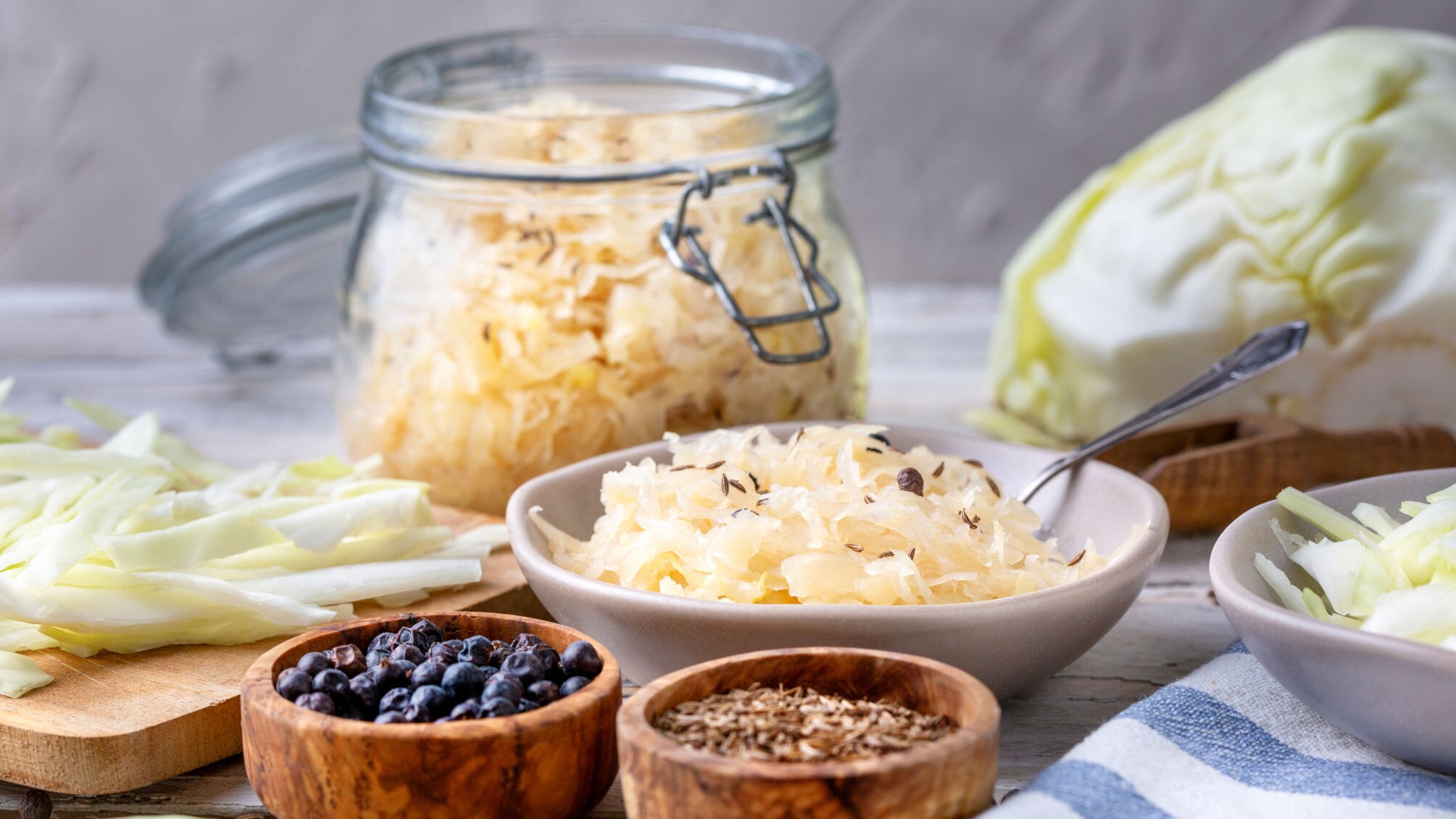 Schüssel mit Sauerkraut, Glas mit Sauerkraut, frischer Weißkohl, Schalen mit Wacholderbeeren und Kümmel auf Holztisch.
