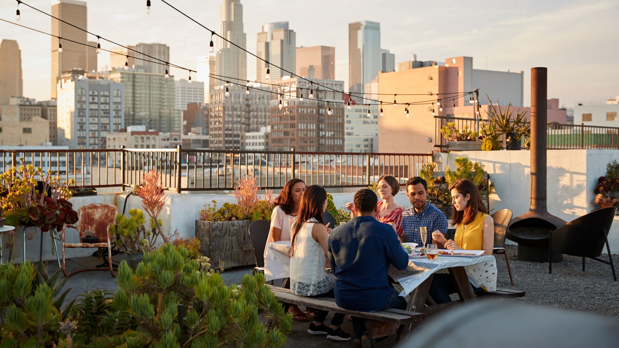 Sechs Personen sitzen beim Essen an einem Tisch auf einer Dachterrasse vor der Skyline einer Großstadt. Sechs Personen sitzen beim Essen an einem Tisch auf einer Dachterrasse vor der Skyline einer Großstadt.