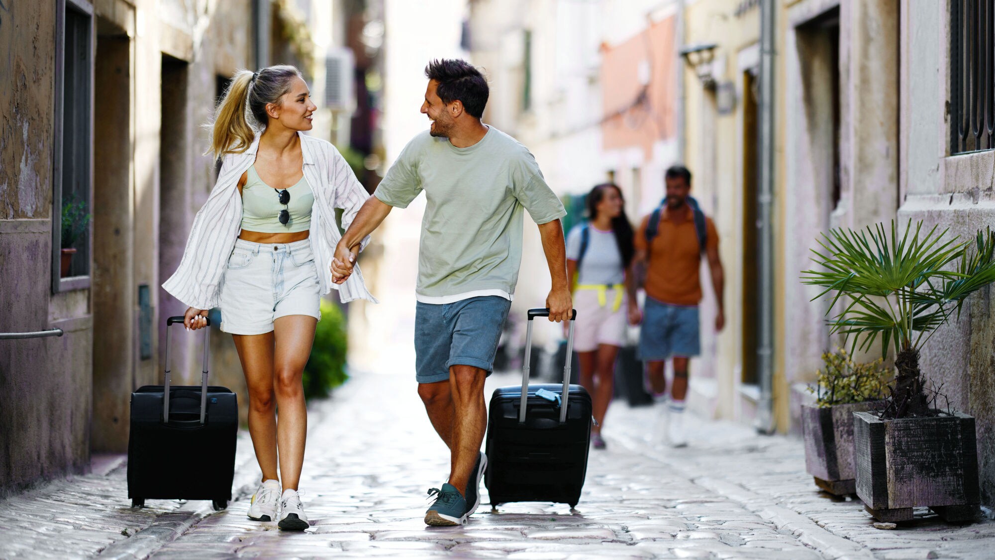 Ein händchenhaltendes Paar in Sommerkleidung geht mit Rollkoffern durch eine Gasse mit Kopfsteinpflaster. Ein händchenhaltendes Paar in Sommerkleidung geht mit Rollkoffern durch eine Gasse mit Kopfsteinpflaster.