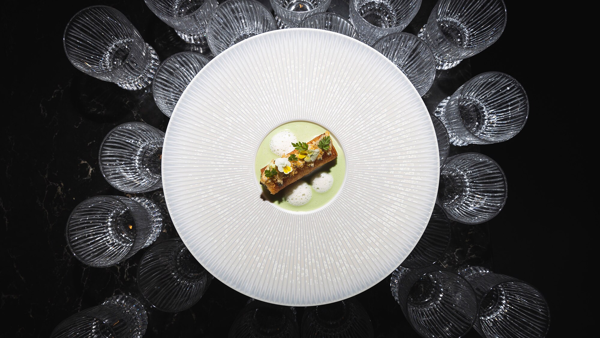 Aufsicht eines elegant angerichteten Gerichtes auf einem großen weißen Teller, der auf mehreren Kristallgläsern auf schwarzem Untergrund steht.