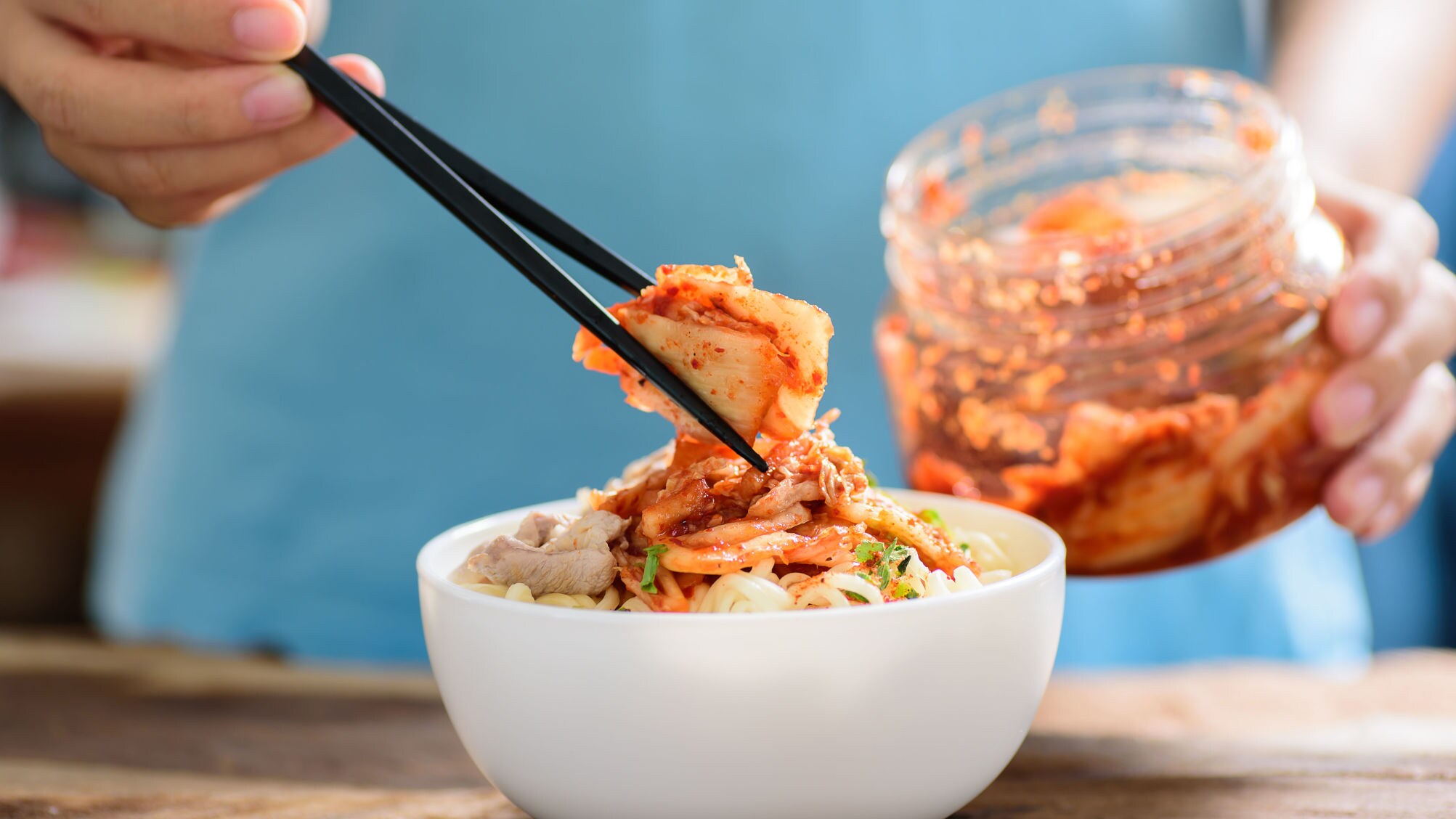 Eine Hand mit Essstäbchen platziert fermentierten Kohl auf einem Gericht in einer weißen Schale, dahinter ein Einmachglas mit Kimchi.