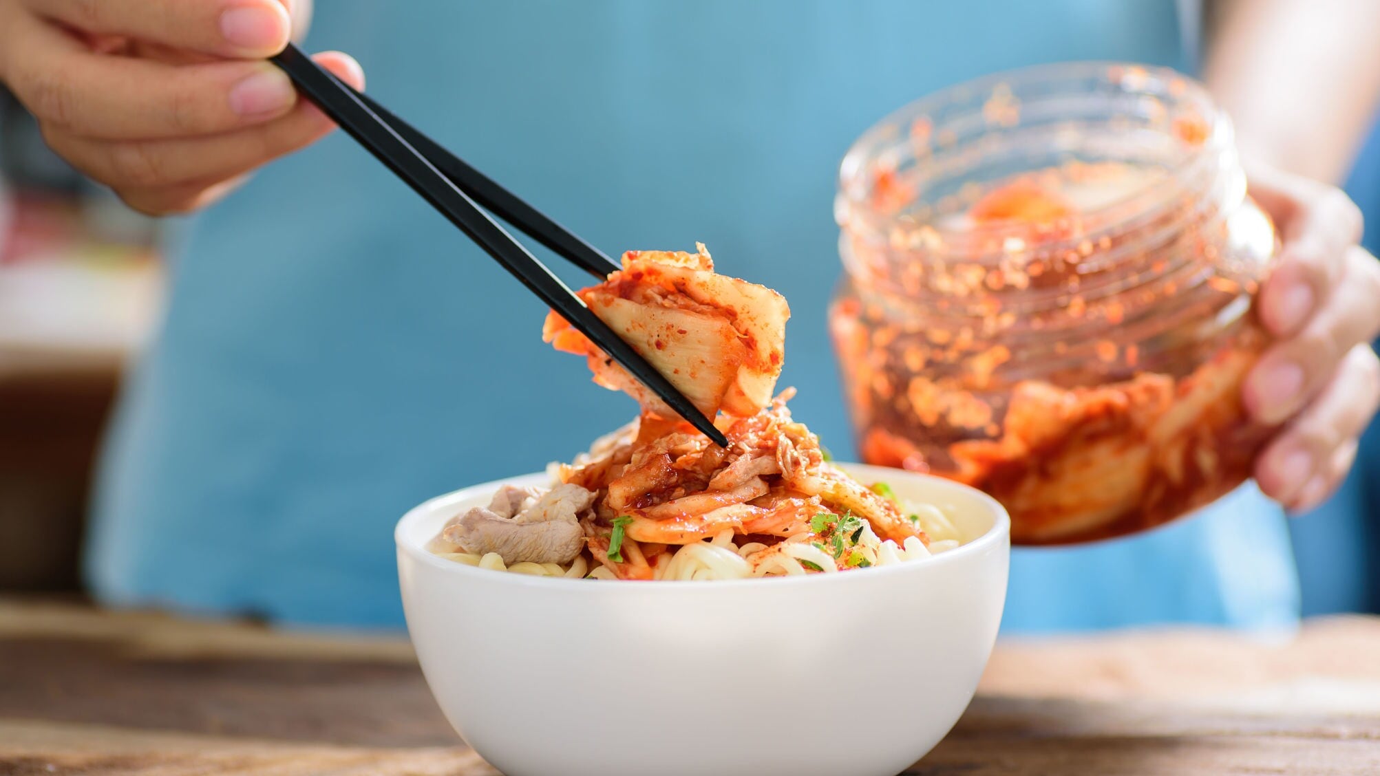 Eine Hand mit Essstäbchen platziert fermentierten Kohl auf einem Gericht in einer weißen Schale, dahinter ein Einmachglas mit Kimchi. Eine Hand mit Essstäbchen platziert fermentierten Kohl auf einem Gericht in einer weißen Schale, dahinter ein Einmachglas mit Kimchi.
