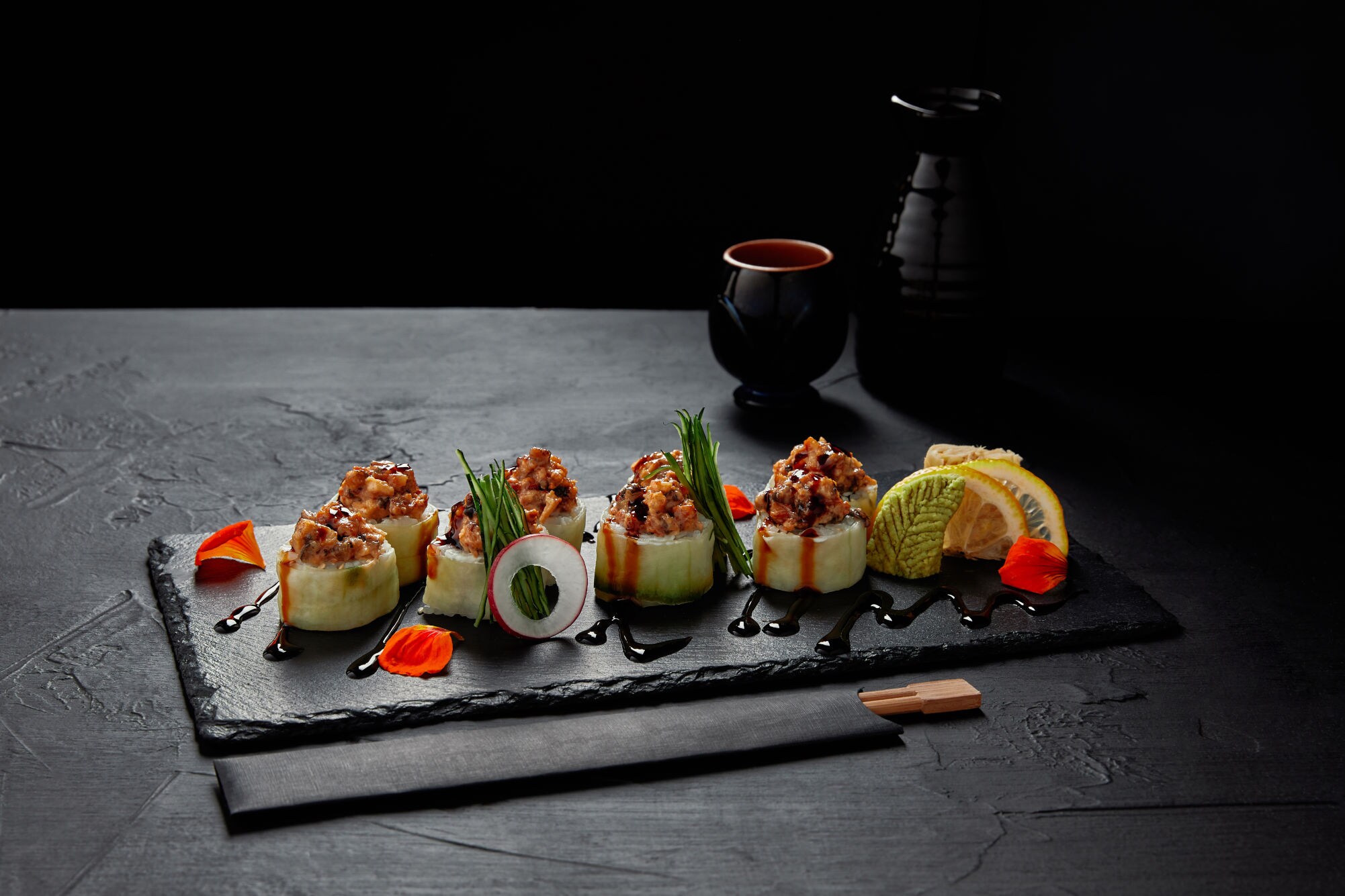 Sushi-Rollen mit rohem Fisch, Gurke, Frühlingszwiebeln und Sojasauce auf schwarzem Schiefer serviert, dekoriert mit Zitronenscheiben und Blütenblättern.