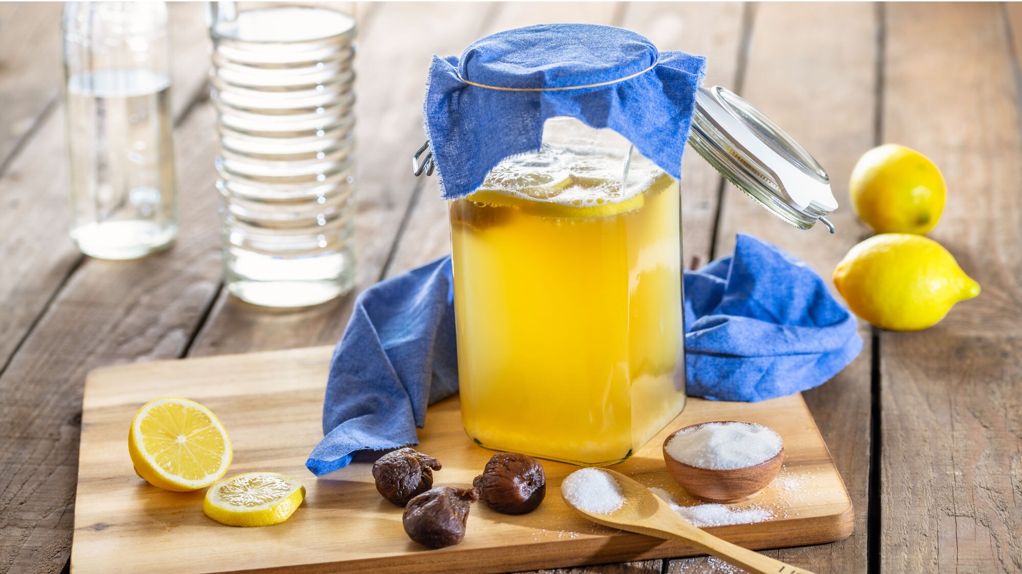 Ein Glasgefäß mit Wasserkefir bedeckt mit einem blauen Tuch, umgeben von Zitronen und weiteren Zutaten auf einem Holzbrett.