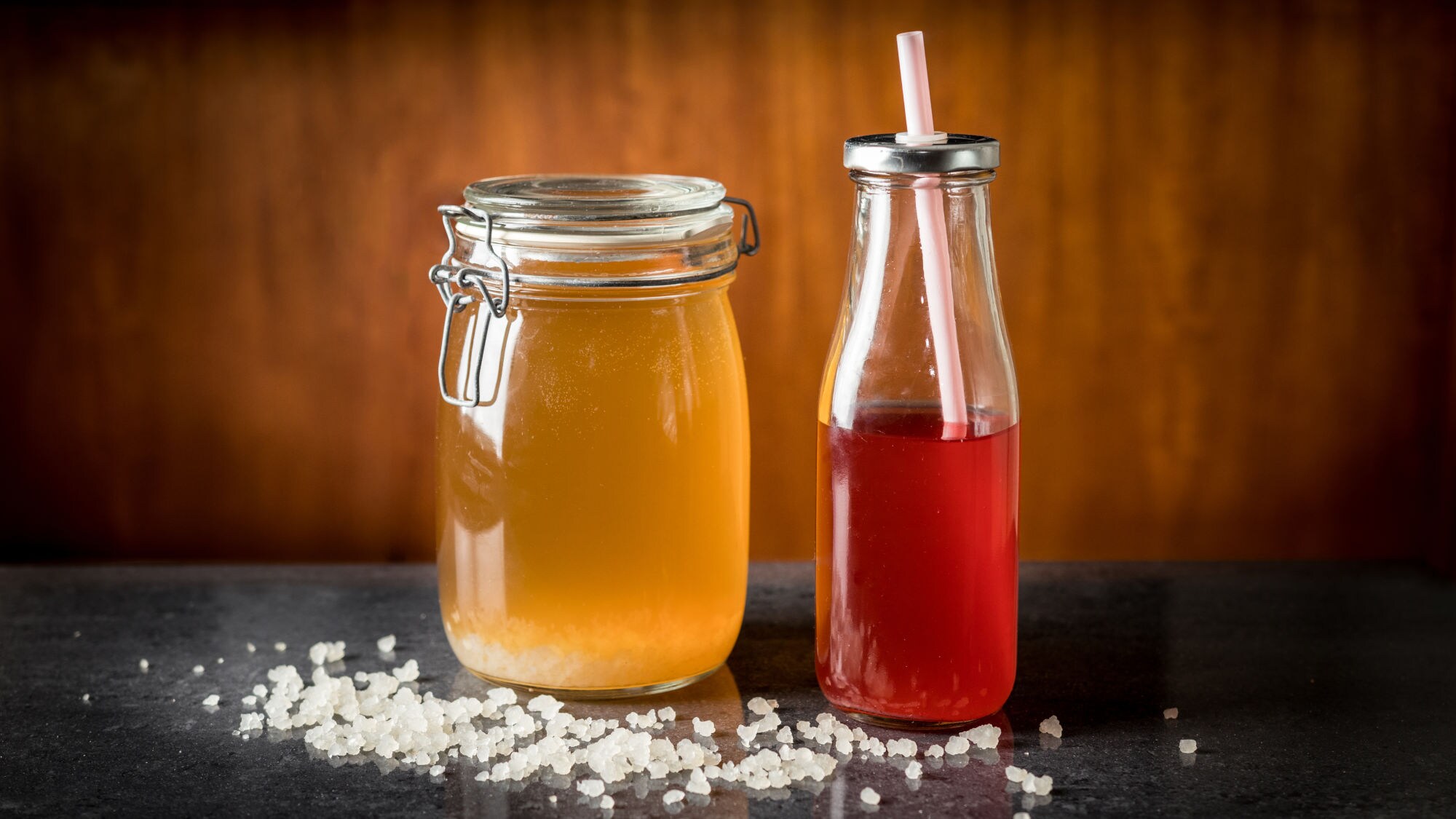 Ein Glas mit gelben und eine Flasche mit rotem Wasserkefir, umgeben von Kristallen auf dunklem Untergrund
