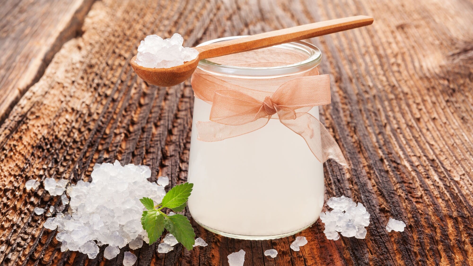 Glas mit milchiger Flüssigkeit und Schleife, umgeben von kristallartigen weißen Körnern auf Holzoberfläche Glas mit milchiger Flüssigkeit und Schleife, umgeben von kristallartigen weißen Körnern auf Holzoberfläche