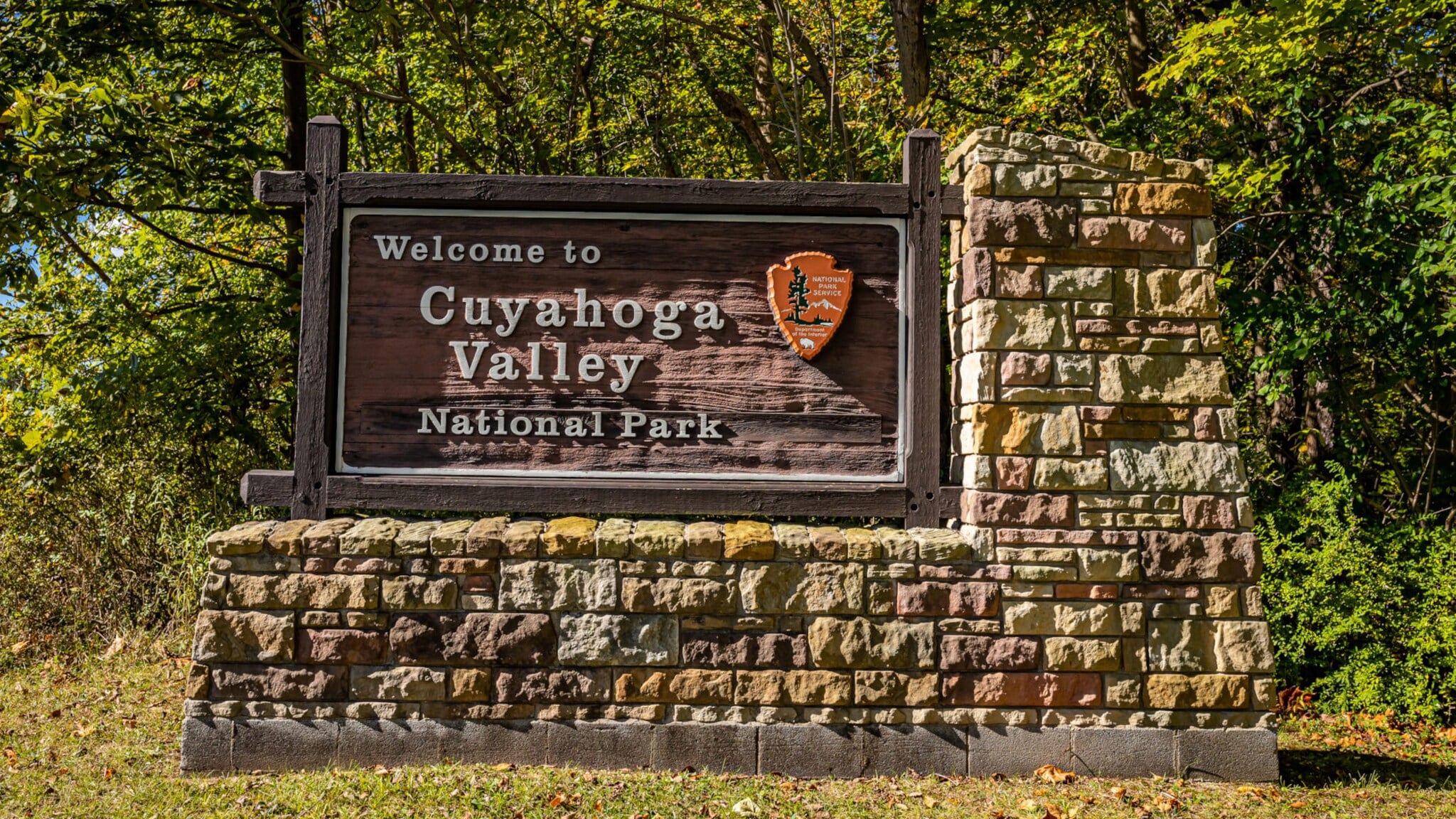 Willkommensschild des Cuyahoga Valley Nationalparks aus Holz auf einem steinernen Sockel, umgeben von Bäumen Willkommensschild des Cuyahoga Valley Nationalparks aus Holz auf einem steinernen Sockel, umgeben von Bäumen