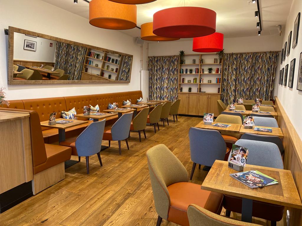 Gastraum mit Holzboden, bunten Stühlen, brauner Polsterbank, großen farbigen Deckenlampen und Bücherregal im Hintergrund.