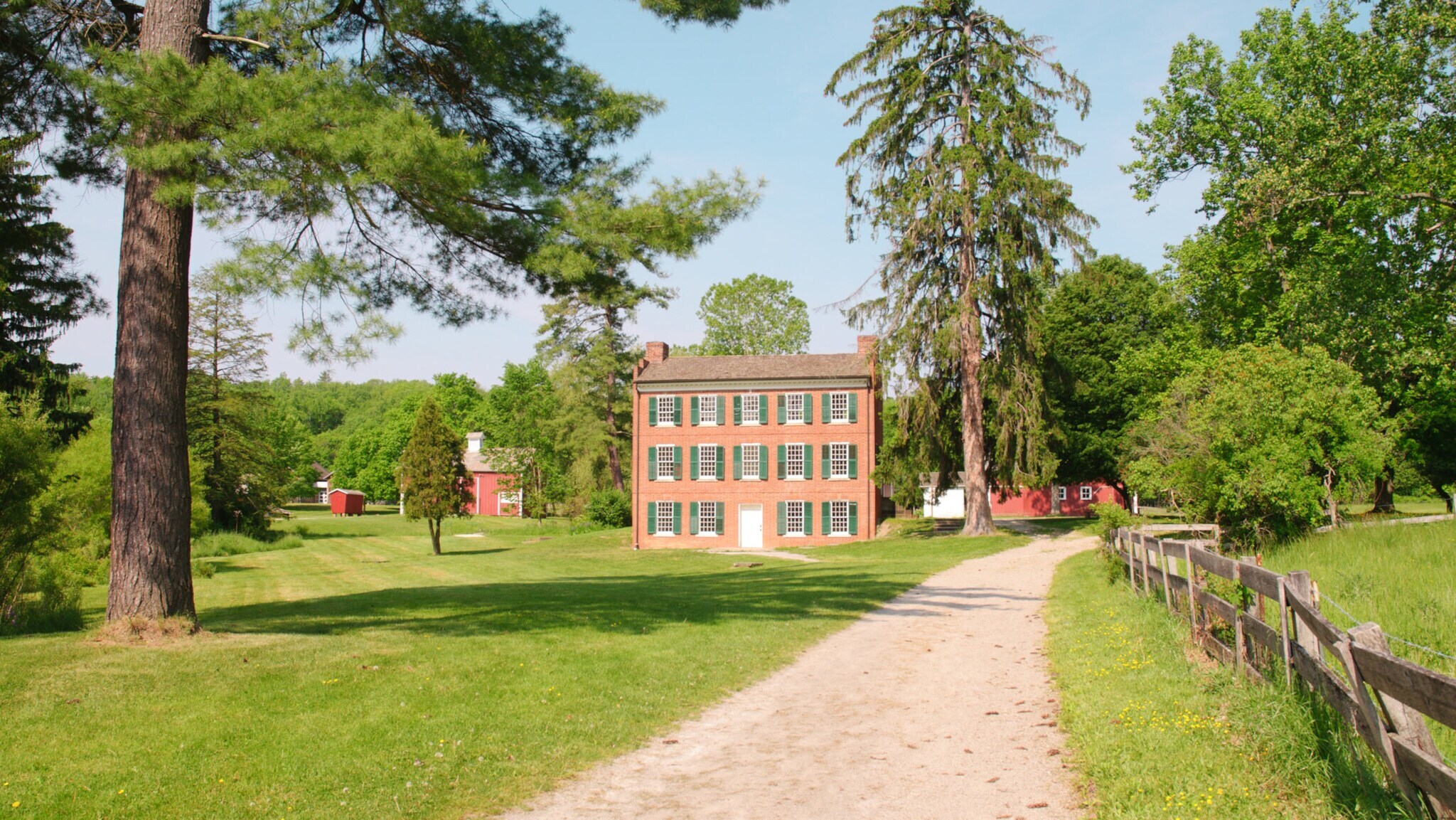Backsteinhaus mit grünen Fensterläden am Ende eines sandigen Wegs, umgeben von großen Bäumen und Wiesen