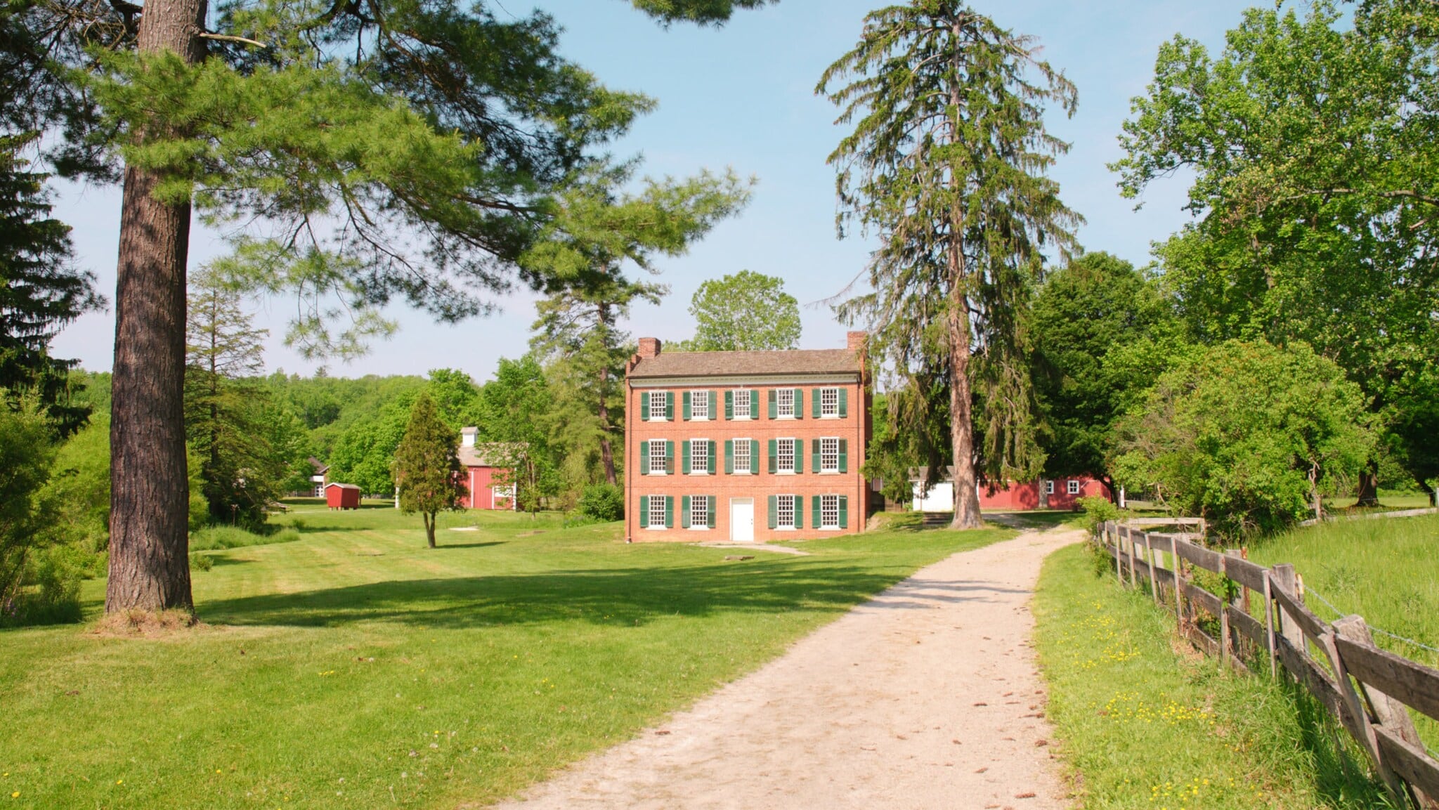 Backsteinhaus mit grünen Fensterläden am Ende eines sandigen Wegs, umgeben von großen Bäumen und Wiesen Backsteinhaus mit grünen Fensterläden am Ende eines sandigen Wegs, umgeben von großen Bäumen und Wiesen