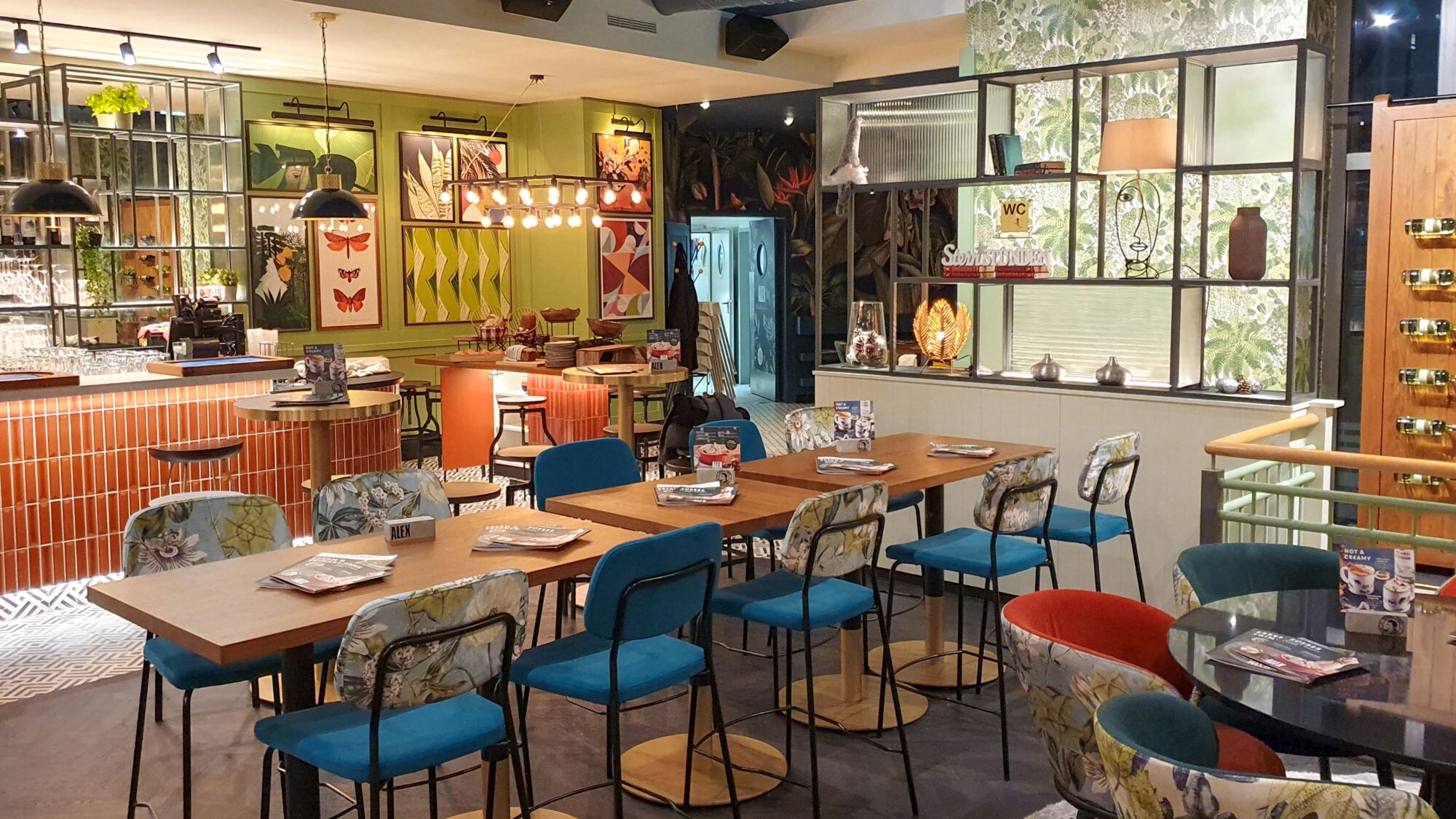 Innenansicht des ALEX in Ludwigshafen, mit Holztischen, bunten Stühlen und dekorativen Bildern an der Wand.