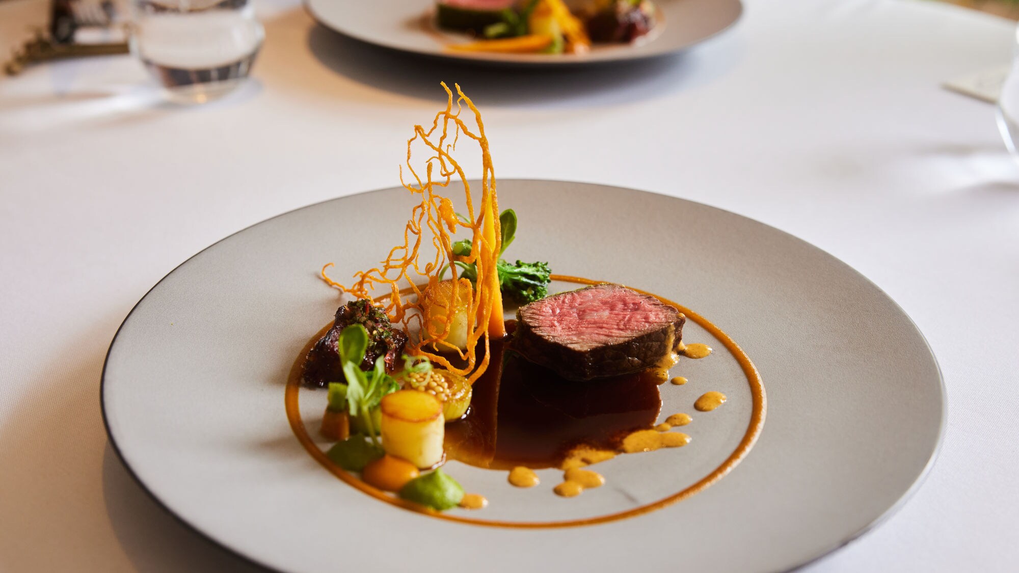 Kunstvoll arrangiertes Gericht mit einem Stück Fleisch auf einem großen weißen Teller auf einem weißen Tisch.