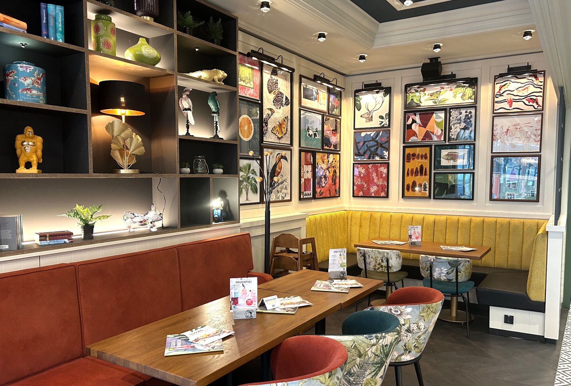 Gemütlicher Gastraum mit gelber Eckbank, bunten Stühlen und zahlreichen farbenfrohen Bildern an den Wänden.