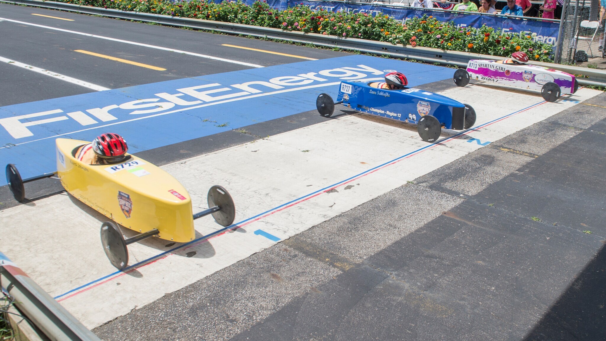 Drei seifenkistenartige Rennwagen in Gelb, Blau und Weiß an der Startlinie eines Rennens auf einer Straße Drei seifenkistenartige Rennwagen in Gelb, Blau und Weiß an der Startlinie eines Rennens auf einer Straße