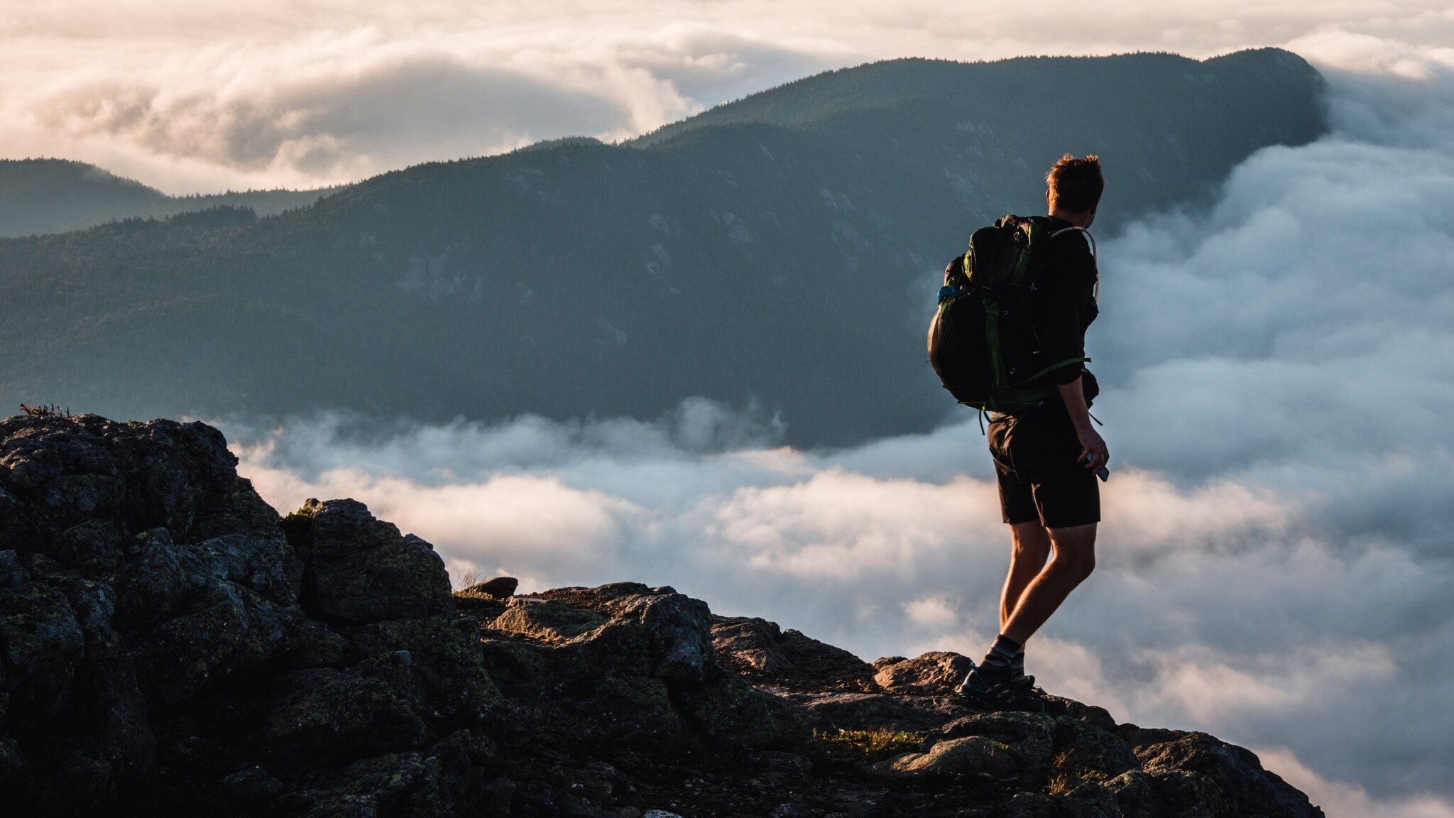 Wanderer mit Rucksack steht auf felsigem Gipfel und blickt auf nebelverhangene Berge bei Sonnenuntergang Wanderer mit Rucksack steht auf felsigem Gipfel und blickt auf nebelverhangene Berge bei Sonnenuntergang