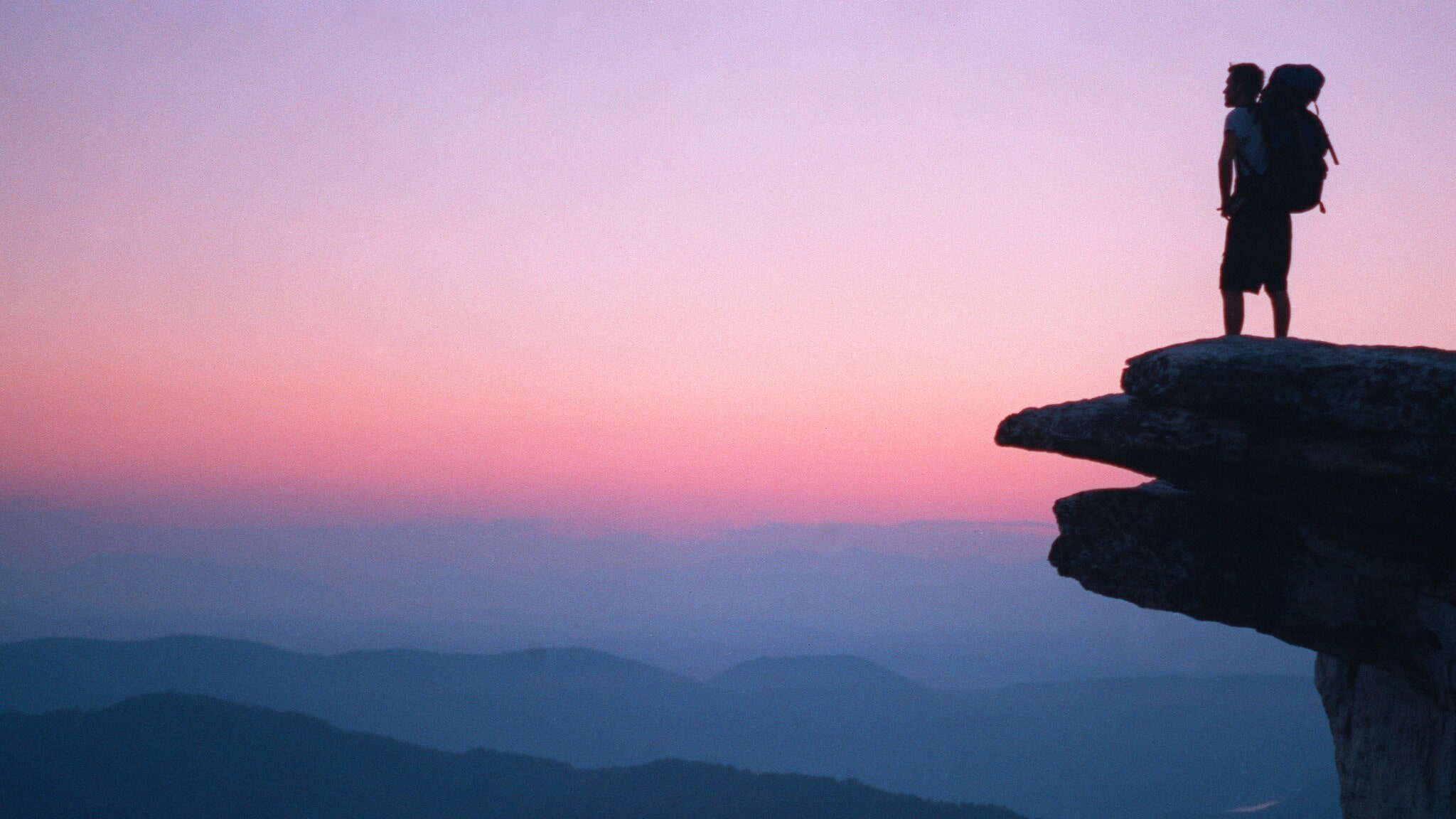 Wanderer mit großem Rucksack steht auf einem Felsen und blickt bei Sonnenuntergang über neblige Berglandschaft Wanderer mit großem Rucksack steht auf einem Felsen und blickt bei Sonnenuntergang über neblige Berglandschaft