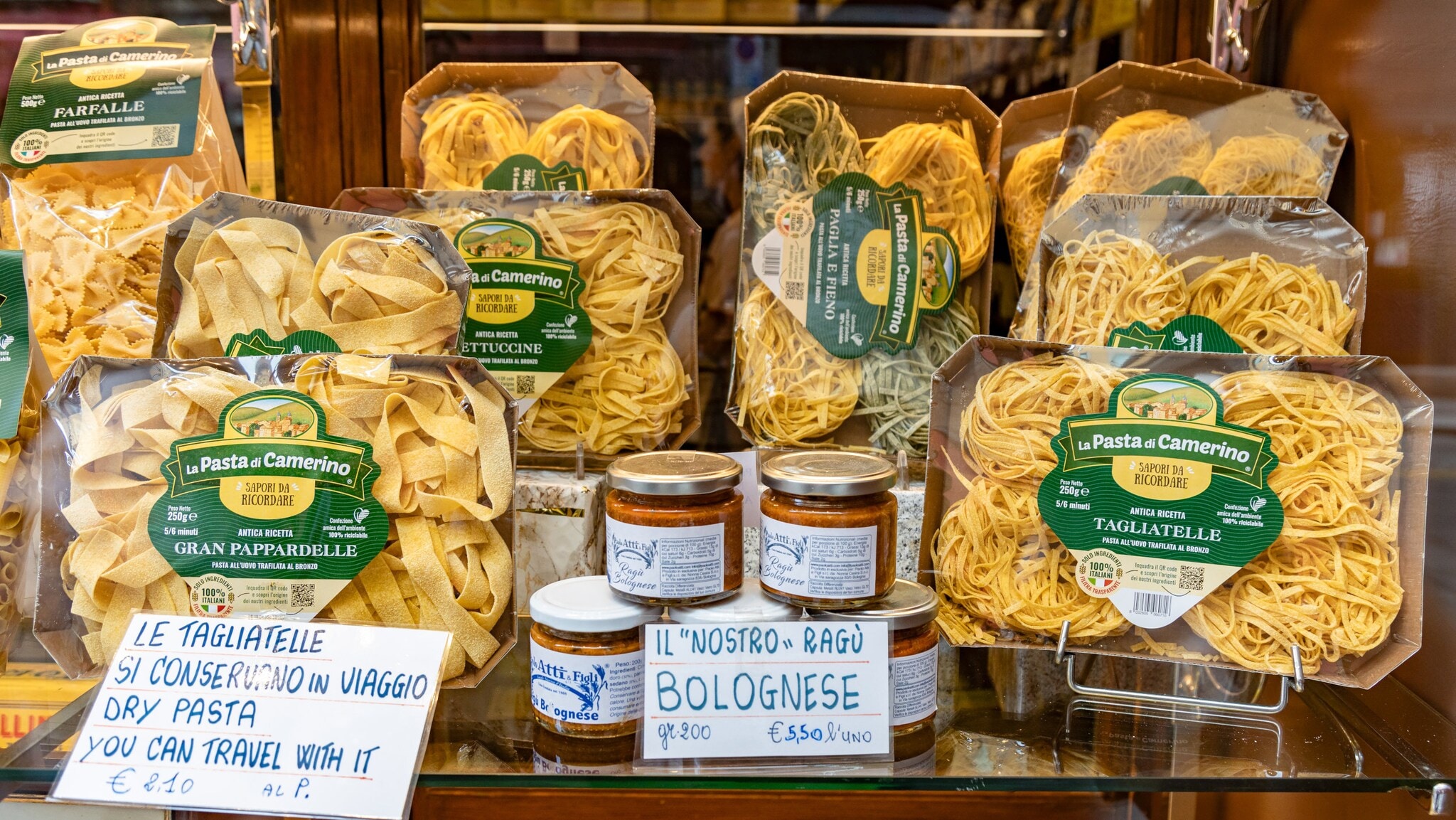 Verschiedene Packungen italienischer Pasta von La Pasta di Camerino und Gläser mit Bolognese-Sauce auf einem Regal, mit handgeschriebenen Preisschildern in Italienisch und Englisch Verschiedene Packungen italienischer Pasta von La Pasta di Camerino und Gläser mit Bolognese-Sauce auf einem Regal, mit handgeschriebenen Preisschildern in Italienisch und Englisch