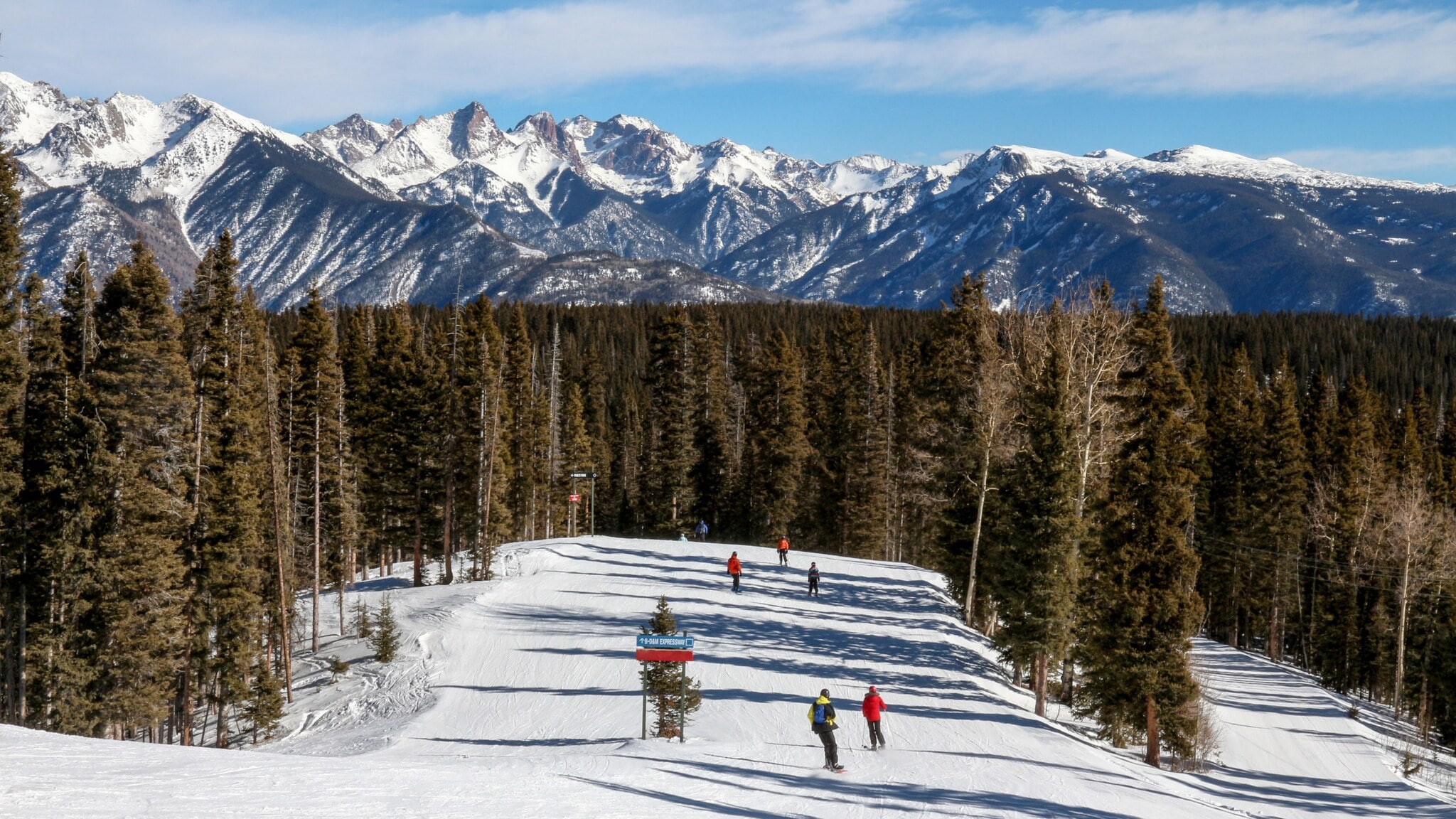 Skifahrer auf einer breiten, schneebedeckten Piste umgeben von hohen Tannen und Bergen im Hintergrund unter blauem Himmel Skifahrer auf einer breiten, schneebedeckten Piste umgeben von hohen Tannen und Bergen im Hintergrund unter blauem Himmel