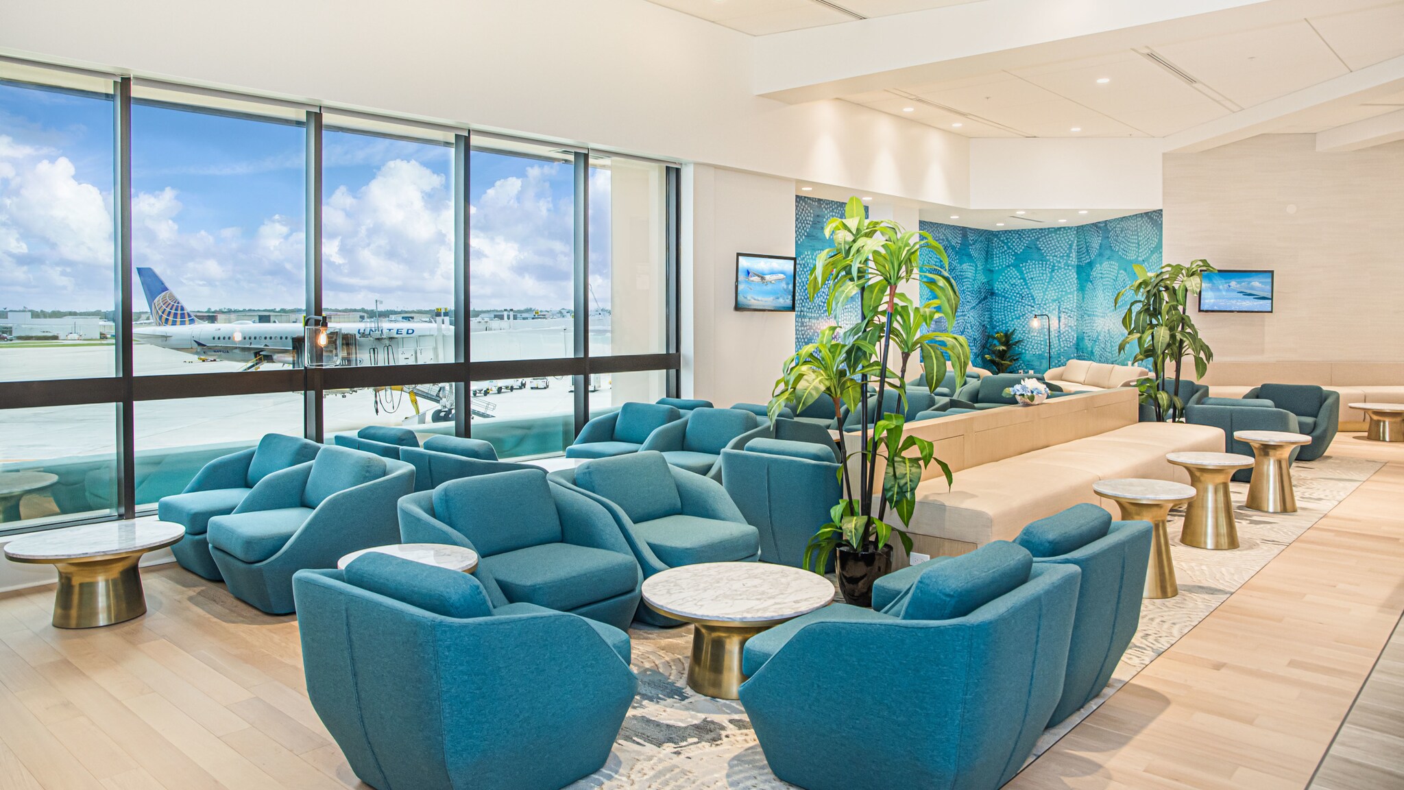 Elegante Flughafen-Lounge mit türkisfarbenen Sesseln und goldfarbenen Tischen mit Blick aufs Rollfeld bei blauem Himmel.