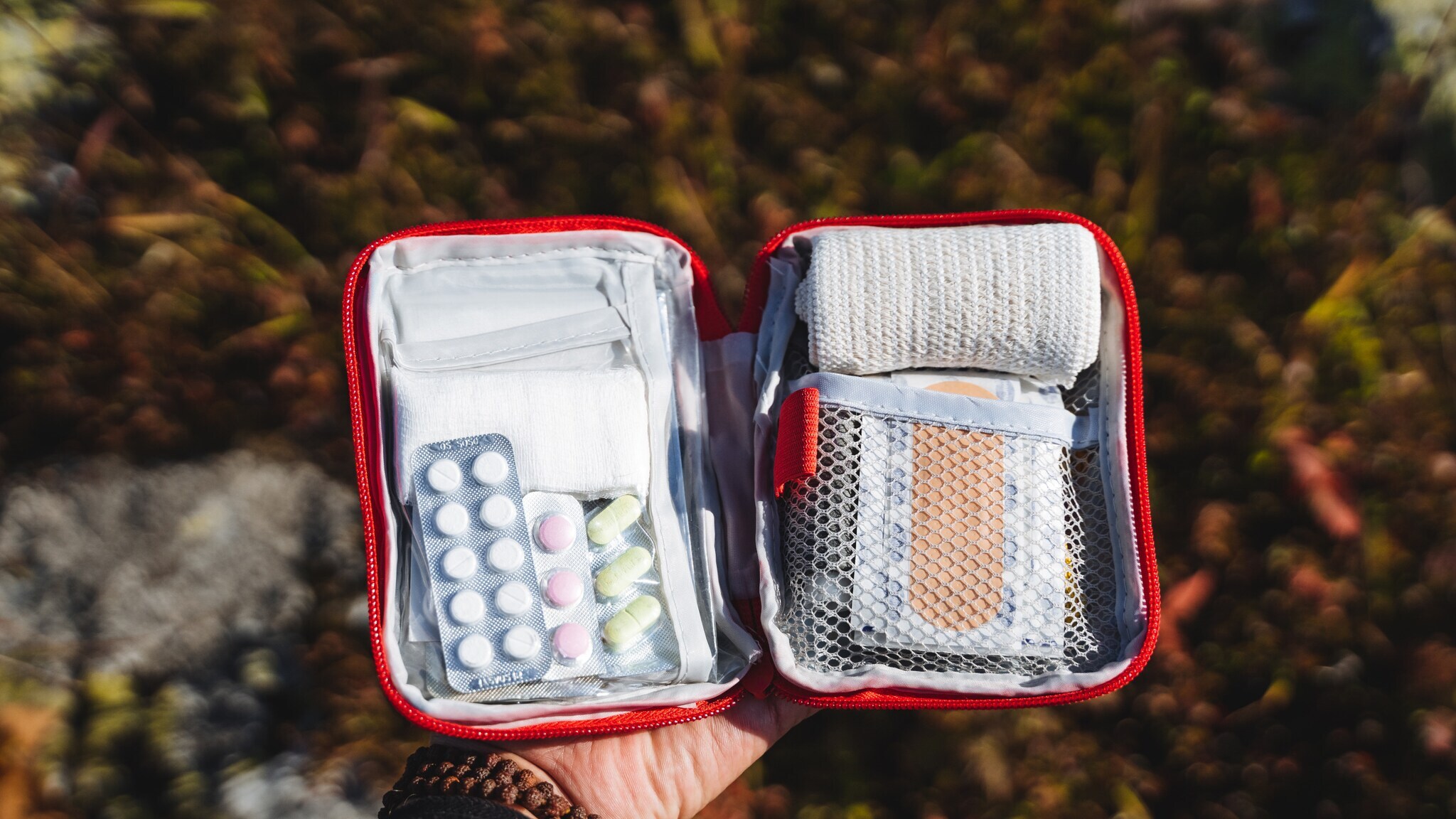 Hand hält eine geöffnete rote Erste-Hilfe-Tasche mit Verbandsmaterial, Pflastern und Medikamenten in Blisterpackungen. Hand hält eine geöffnete rote Erste-Hilfe-Tasche mit Verbandsmaterial, Pflastern und Medikamenten in Blisterpackungen.