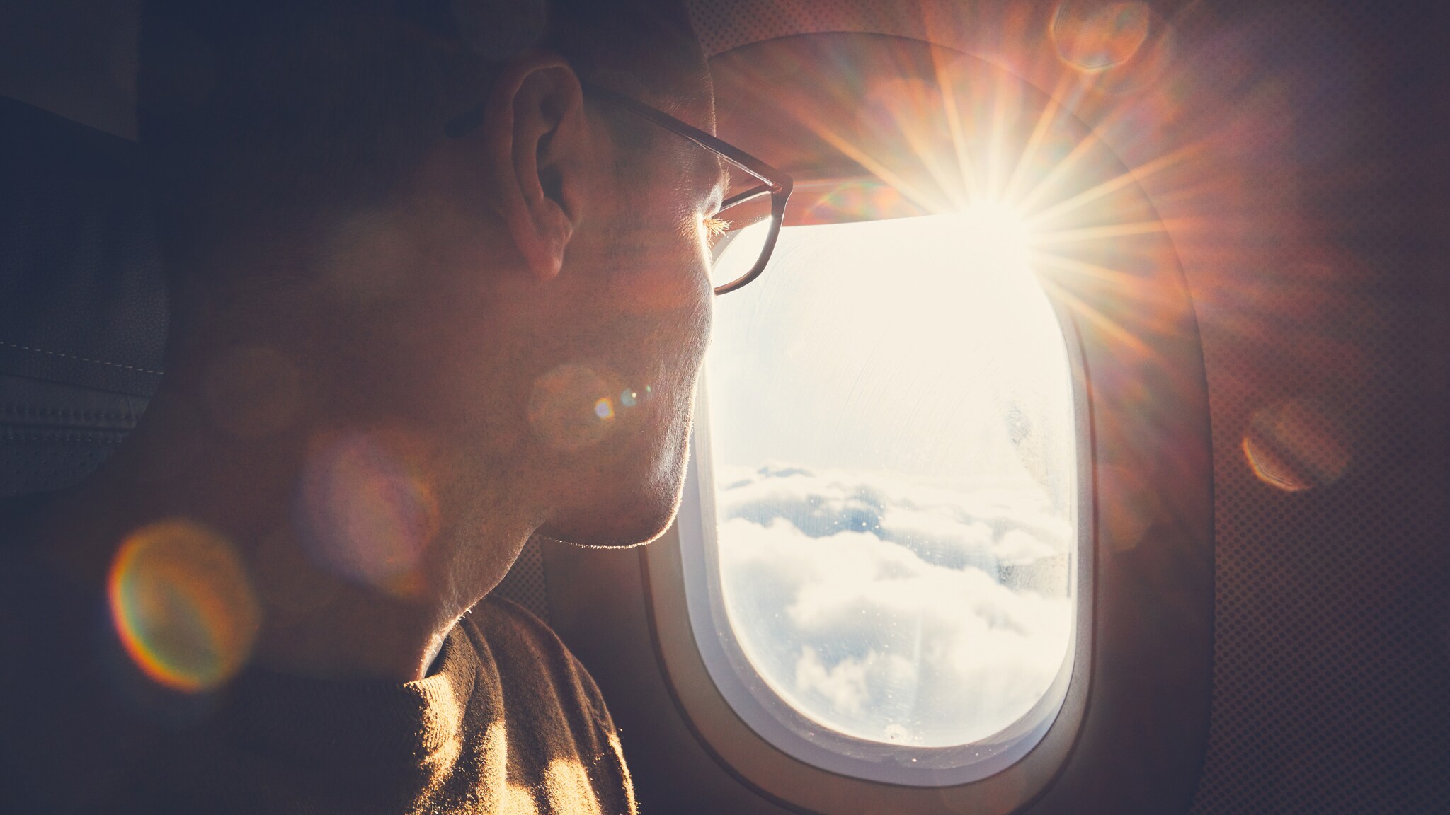 Mann mit Brille blickt aus dem Flugzeugfenster auf die Sonne und Wolken
