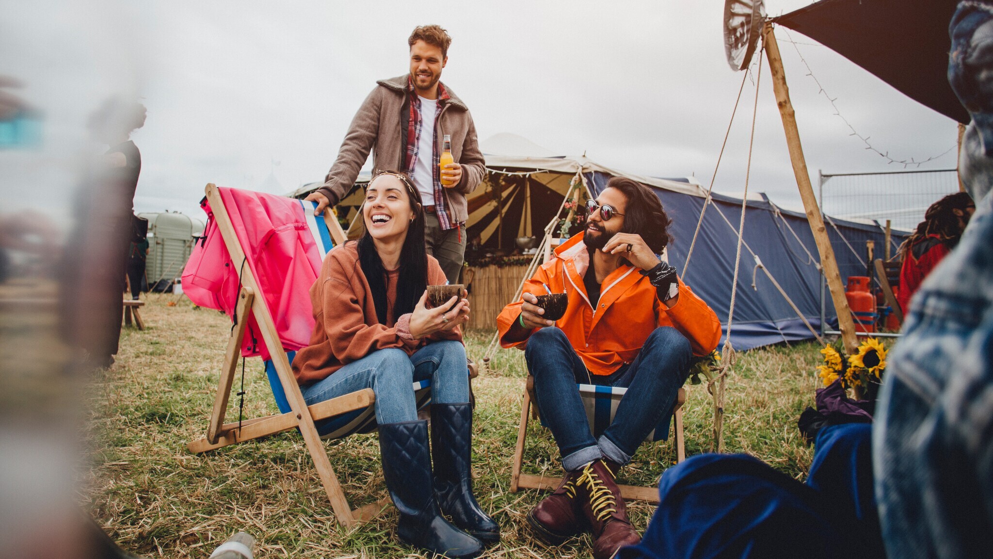 Drei Personen sitzen und stehen entspannt auf einem Festivalgelände mit Zelten im Hintergrund, lachen und trinken Getränke.