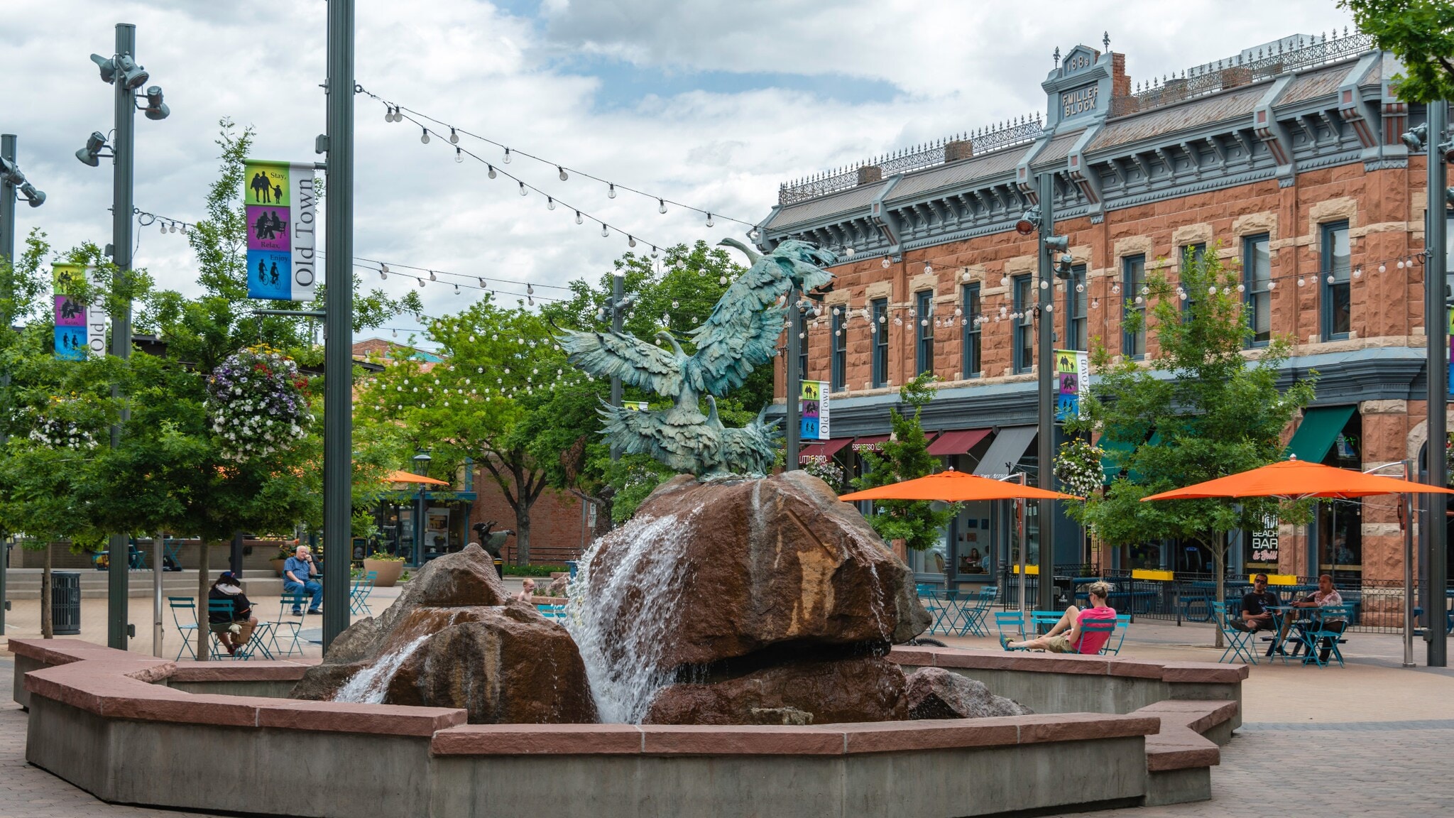 Brunnen mit Adlerstatue auf einem Felsen in einem belebten Platz mit historischen Gebäuden und Sitzbereichen mit orangenen Sonnenschirmen Brunnen mit Adlerstatue auf einem Felsen in einem belebten Platz mit historischen Gebäuden und Sitzbereichen mit orangenen Sonnenschirmen