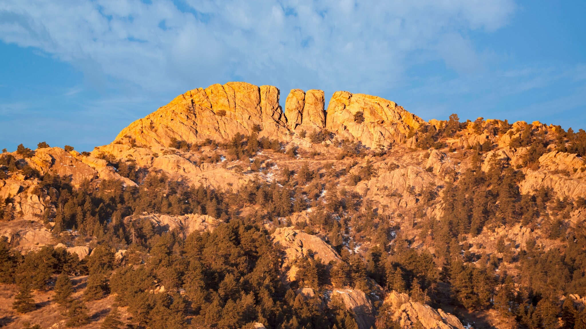 Felsiger Berg mit orangefarbenem Sonnenlicht und bewaldeten Hängen unter blauem Himmel mit Wolken Felsiger Berg mit orangefarbenem Sonnenlicht und bewaldeten Hängen unter blauem Himmel mit Wolken