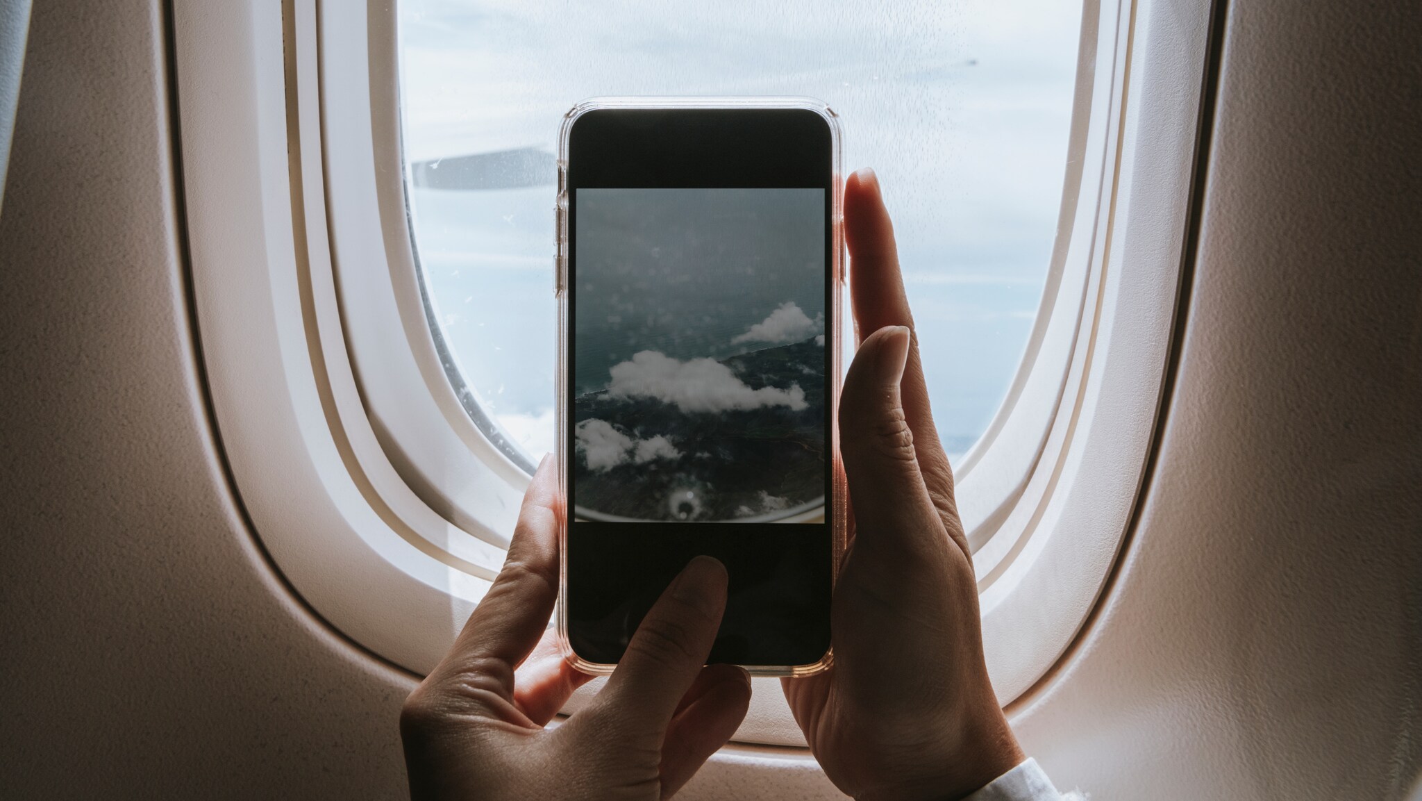 Hände halten ein Smartphone vor einem Flugzeugfenster, das den Himmel mit Wolken zeigt. Hände halten ein Smartphone vor einem Flugzeugfenster, das den Himmel mit Wolken zeigt.