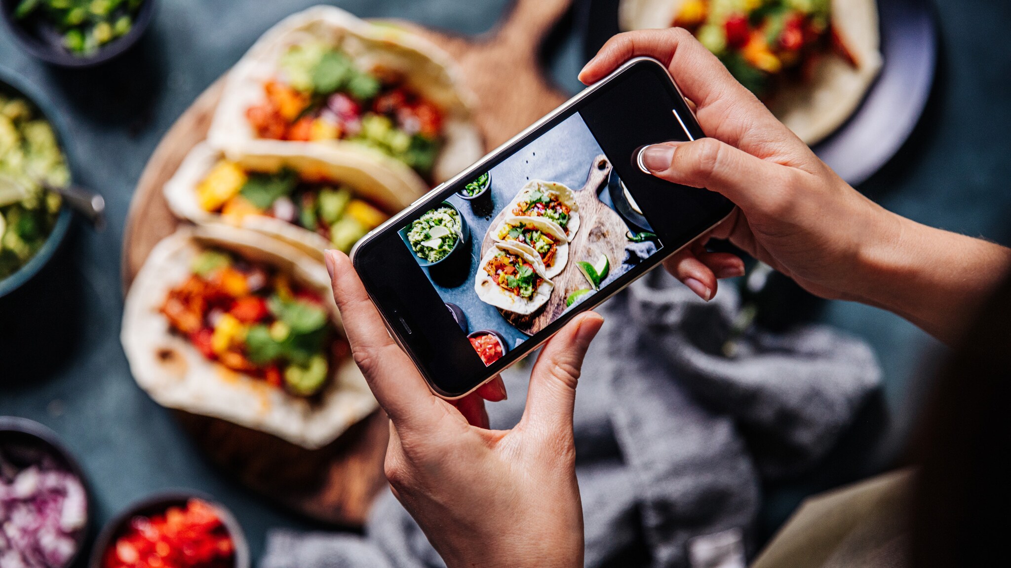 Person fotografiert mit Smartphone drei gefüllte Tacos auf einem Holzbrett, umgeben von kleinen Schalen mit Salsa und Guacamole Person fotografiert mit Smartphone drei gefüllte Tacos auf einem Holzbrett, umgeben von kleinen Schalen mit Salsa und Guacamole