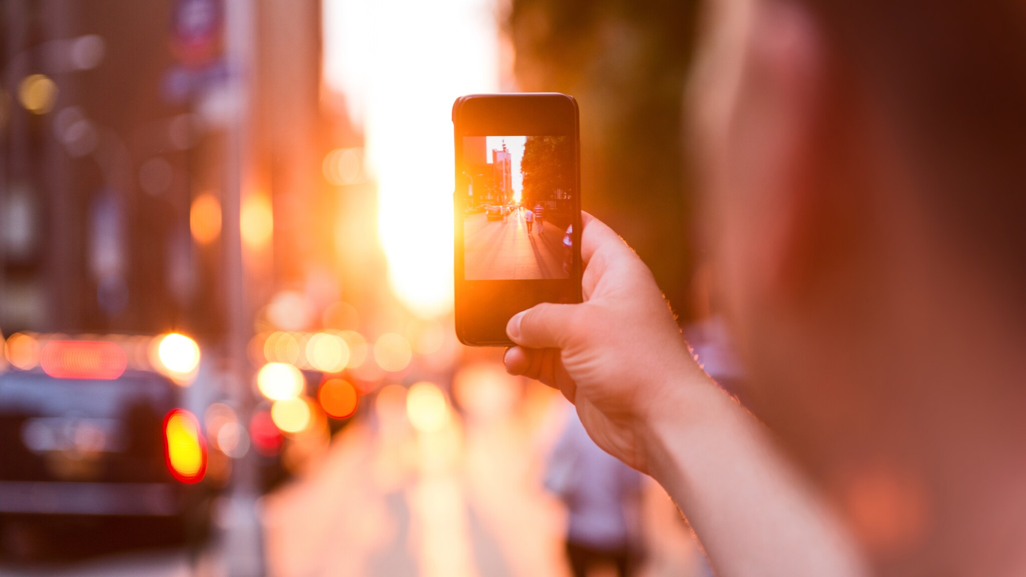Person hält Smartphone und fotografiert eine belebte Straße bei Sonnenuntergang mit verschwommenem Hintergrund