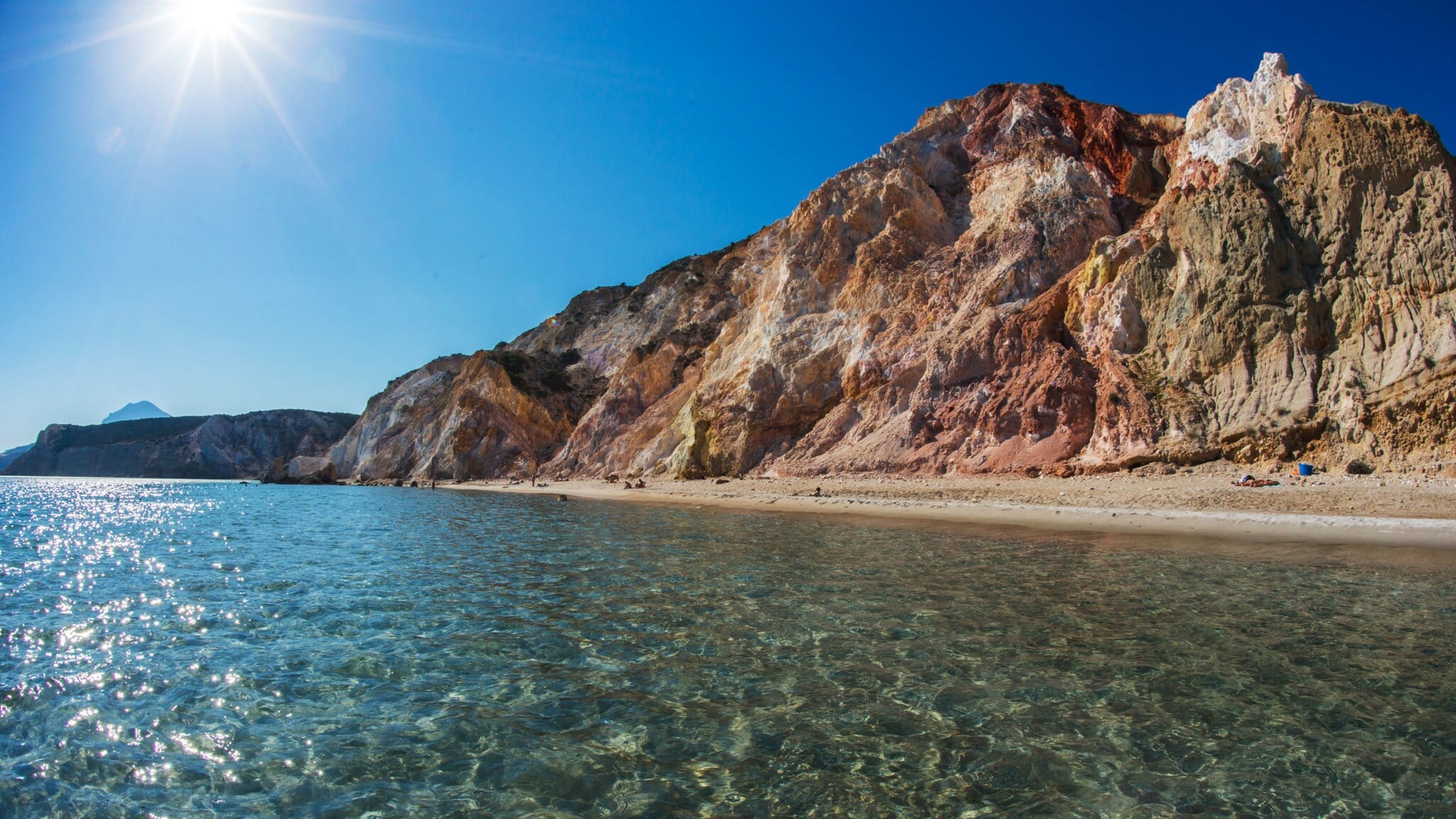 Kleiner Sandstrand an kristallklarem Wasser vor einer Felswand mit unterschiedlichen Gesteinsfarben bei Sonnenschein. Kleiner Sandstrand an kristallklarem Wasser vor einer Felswand mit unterschiedlichen Gesteinsfarben bei Sonnenschein.