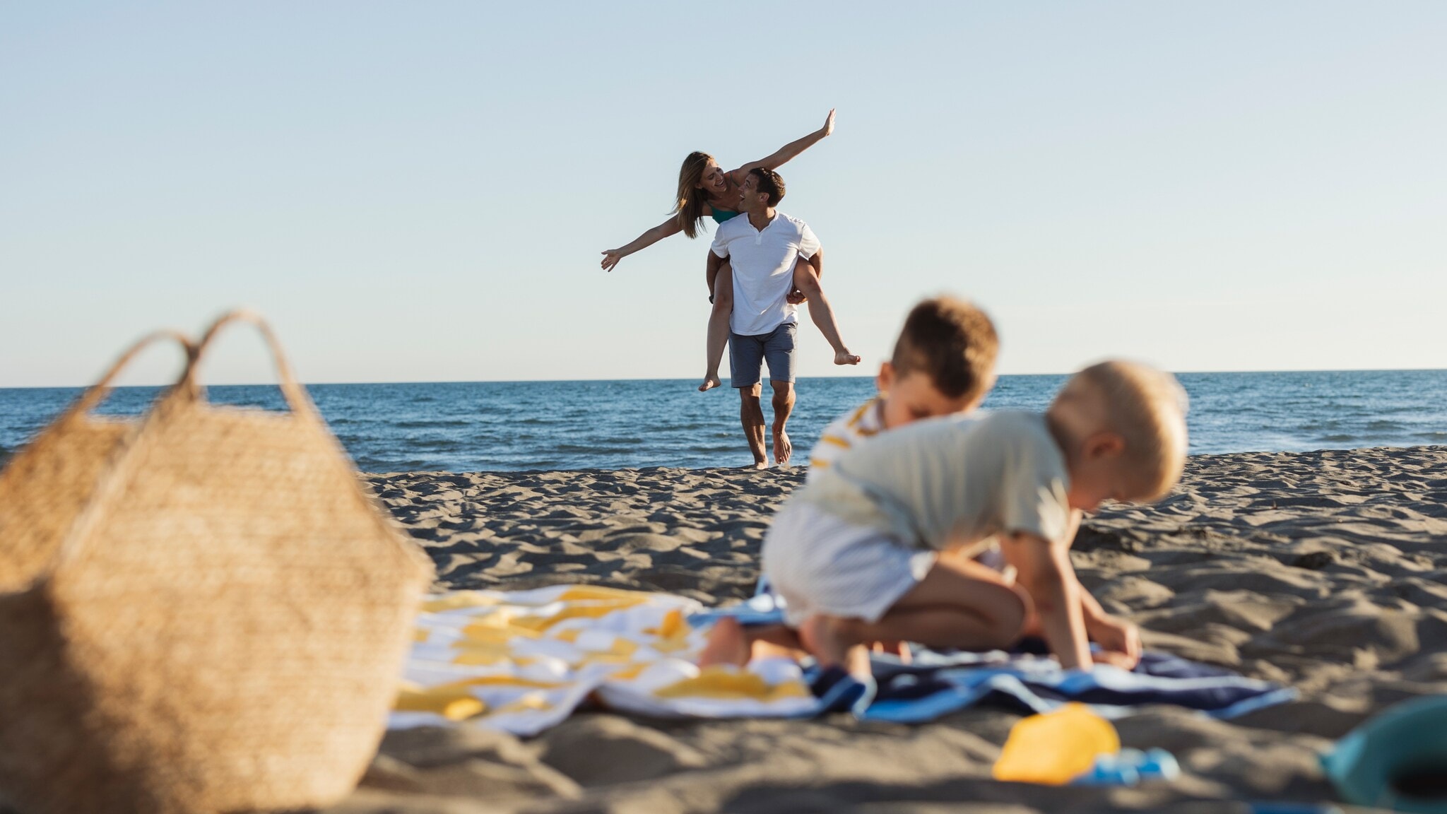 Zwei spielende Kinder auf einem Strandtuch am Strand, im Hintergrund trägt ein Mann eine Frau auf dem Rücken am Meer Zwei spielende Kinder auf einem Strandtuch am Strand, im Hintergrund trägt ein Mann eine Frau auf dem Rücken am Meer