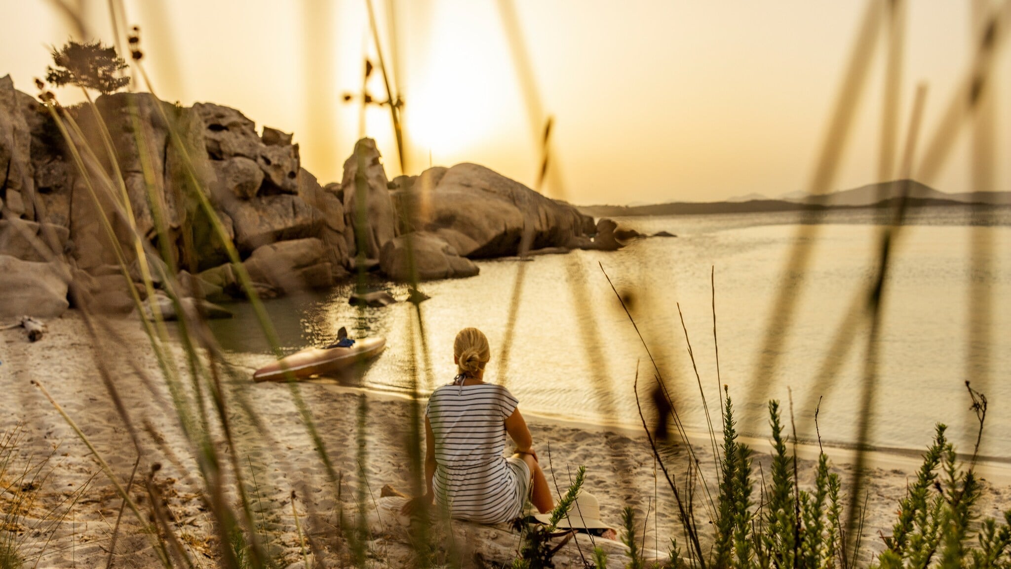 Person sitzt am Ufer eines Sees oder Meeres bei Sonnenuntergang, umgeben von Gras und Felsen Person sitzt am Ufer eines Sees oder Meeres bei Sonnenuntergang, umgeben von Gras und Felsen