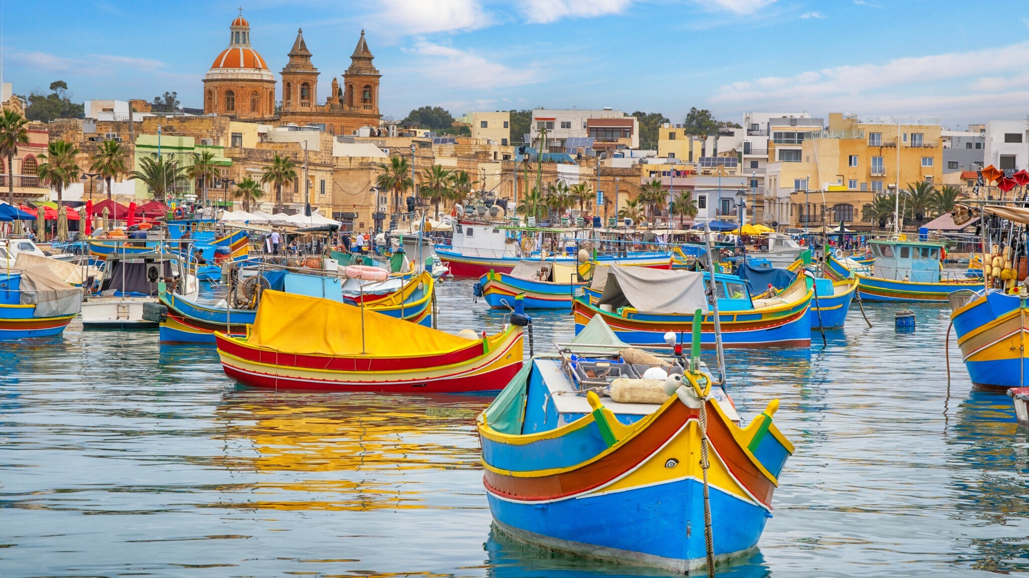 Bunte traditionelle Fischerboote im Hafen von Marsaxlokk, Malta, mit Kirche und Gebäuden im Hintergrund.