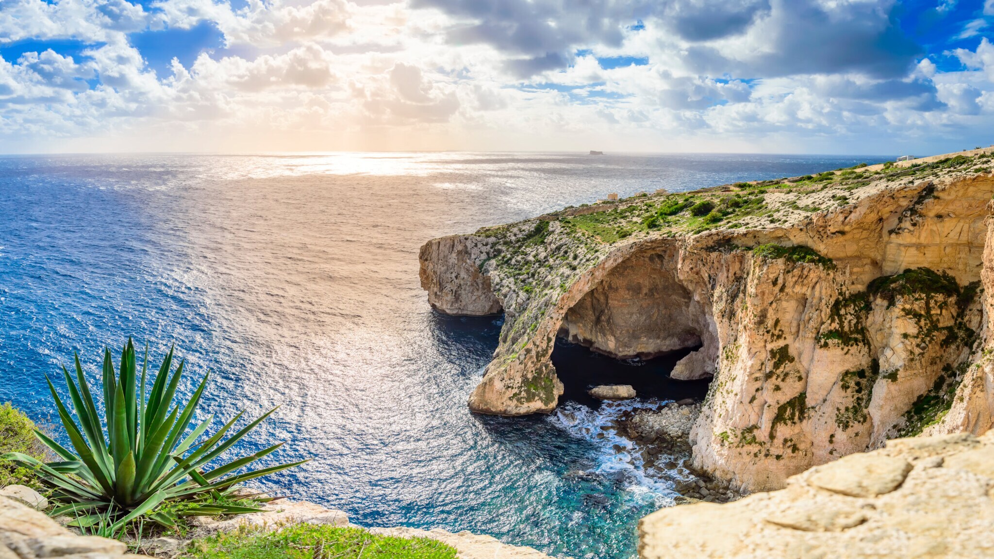 Felsformation mit natürlichem Bogen über blauem Meer unter bewölktem Himmel bei Sonnenuntergang.