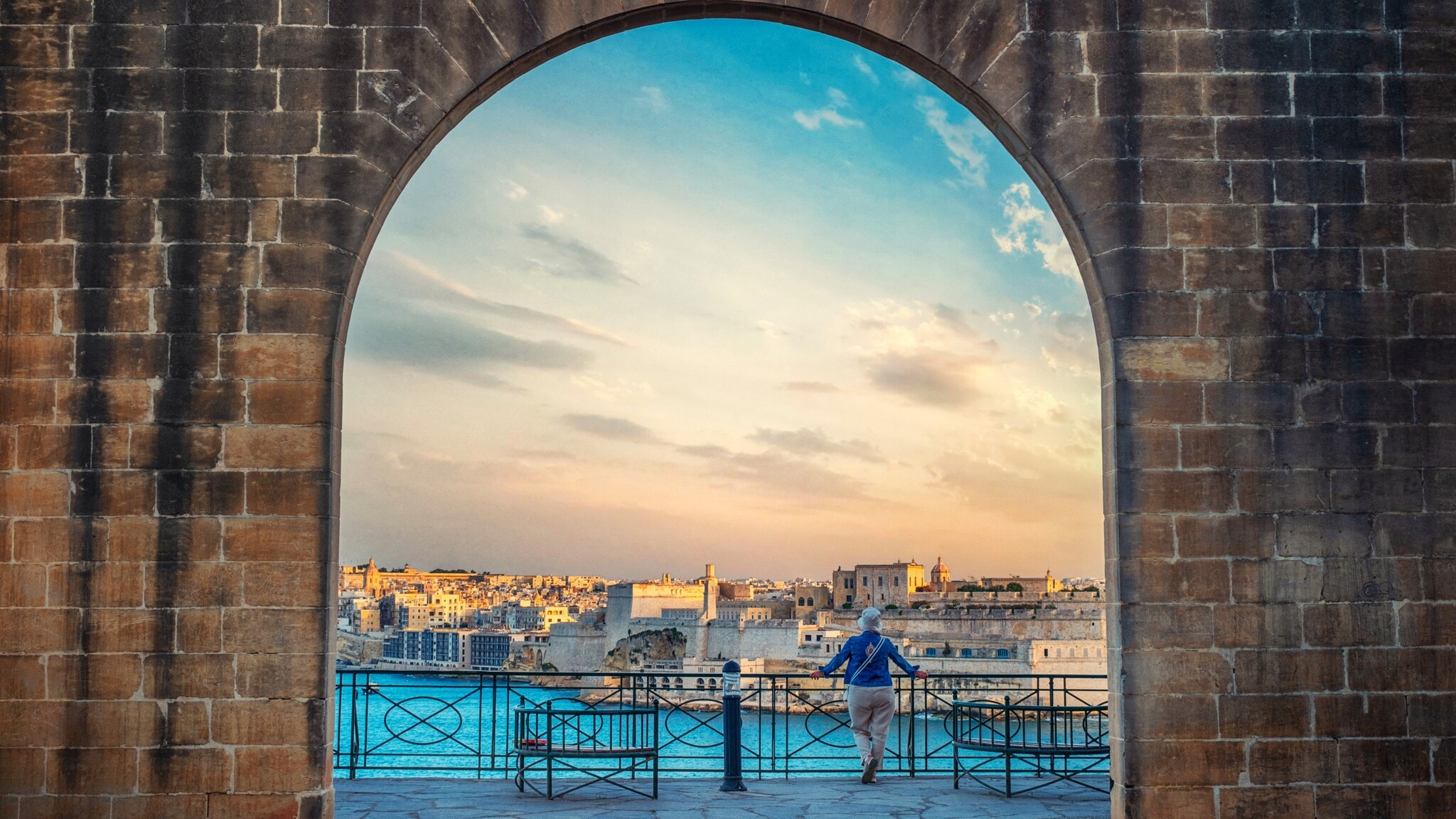 Blick durch einen großen Steinbogen auf eine Stadt am Wasser bei Sonnenuntergang, eine Person lehnt am Geländer Blick durch einen großen Steinbogen auf eine Stadt am Wasser bei Sonnenuntergang, eine Person lehnt am Geländer