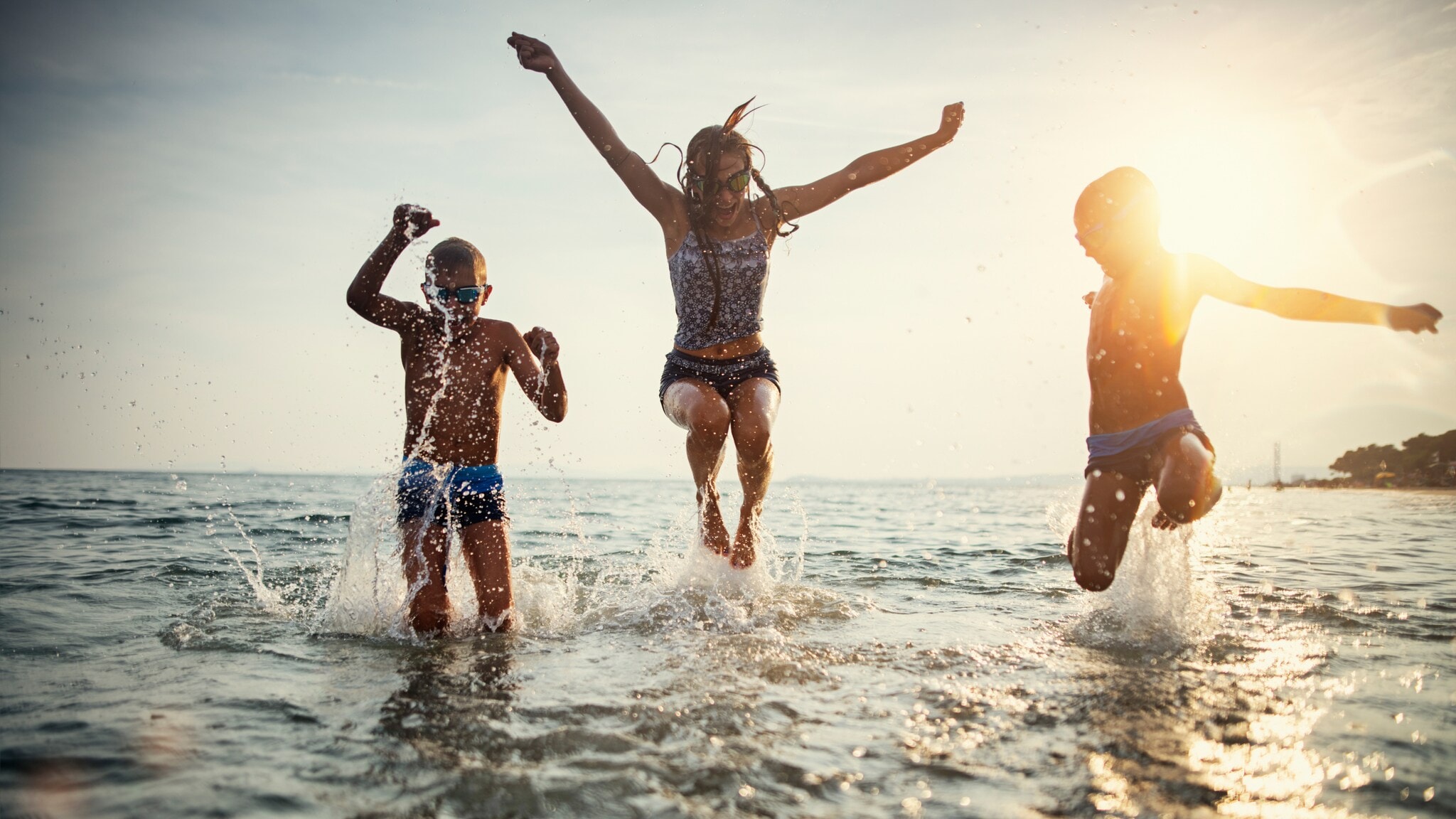 Drei Kinder springen lachend im flachen Wasser am Strand bei Sonnenuntergang Drei Kinder springen lachend im flachen Wasser am Strand bei Sonnenuntergang