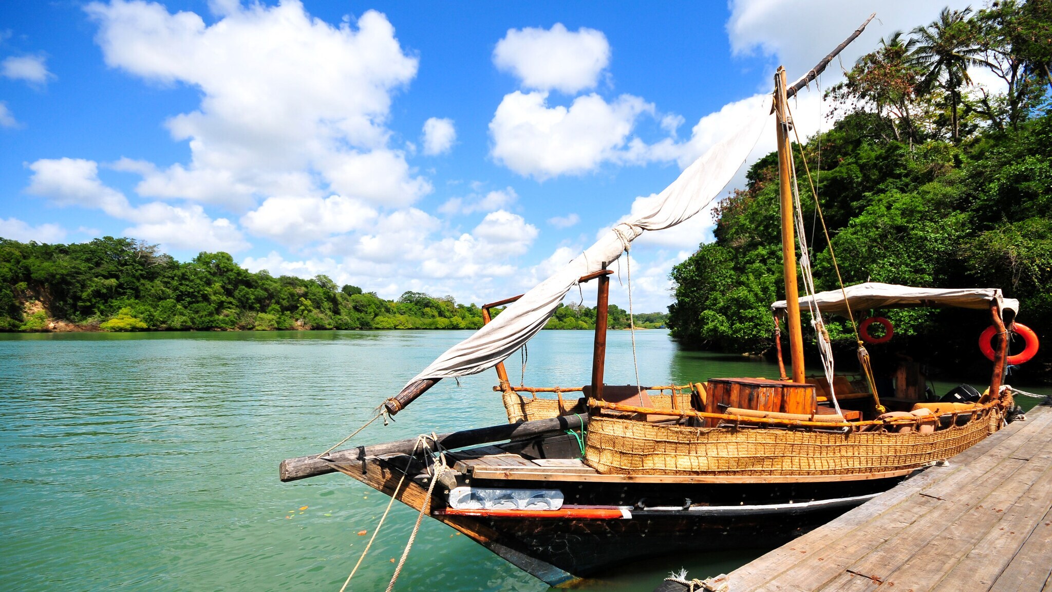 Traditionelles Holzsegelboot mit gefalteten weißen Segeln am Ufer eines ruhigen Flusses, umgeben von grünen Bäumen unter blauem Himmel mit Wolken