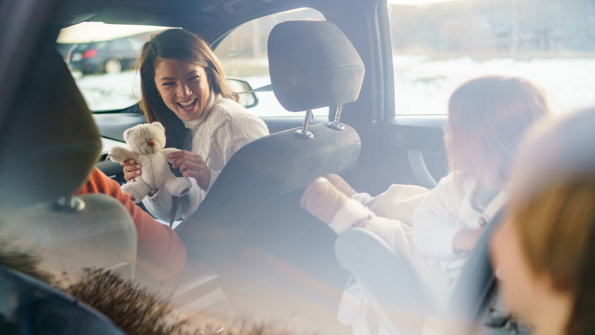 Frau im Auto hält einen weißen Teddybär und lacht, während zwei Kinder auf den Rücksitzen sitzen